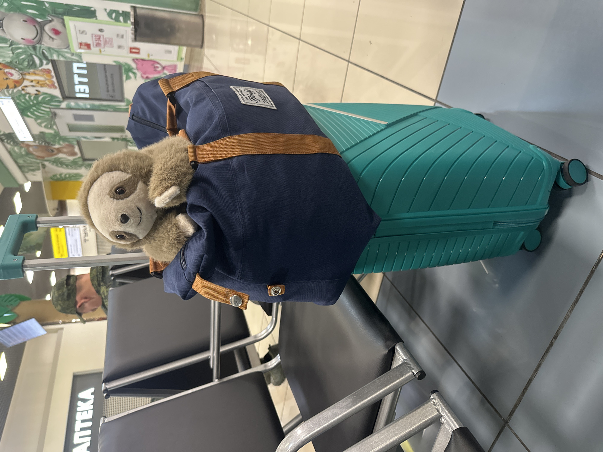 The image depicts a turquoise suitcase with a navy blue duffel bag resting on top of it. A stuffed sloth toy is peeking out of the duffel bag. The luggage is situated in what appears to be an airport waiting area, with rows of dark gray chairs visible to the left. In the background, there's a wall adorned with cartoonish jungle animal murals. A person wearing a military uniform and a camouflage cap is seated in one of the chairs in the distance, facing away from the camera. Signs with Russian text are also visible on the walls, including one that reads "АПТЕКА" (APTEKA, meaning "pharmacy"). The flooring is tiled in shades of gray and beige. The presence of airport seating and signage suggests the location is likely Artyom Airport (VKO) in Artyom, Russia, given the context of the prompt.