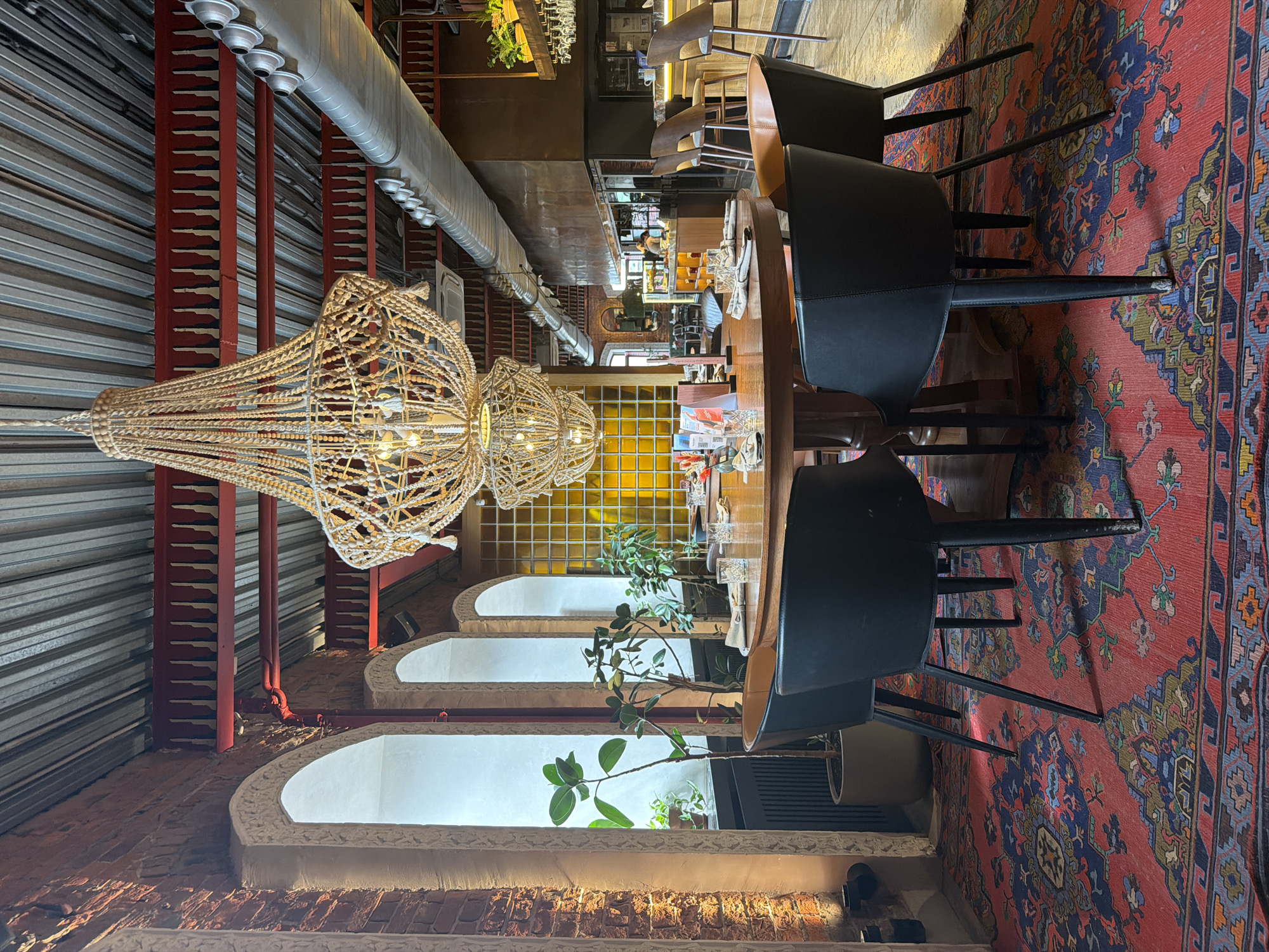 The image shows the interior of a restaurant with a round wooden table set for a meal. Four black chairs surround the table, and the floor is covered with a large, colorful Persian rug. Two ornate chandeliers hang from the ceiling, which is constructed with corrugated metal and exposed red beams. To the left, a brick wall features arched windows with light filtering through, and potted plants are placed in front of them. In the background, a bar area is visible with stools and a counter. The overall ambiance suggests a stylish and contemporary dining space.