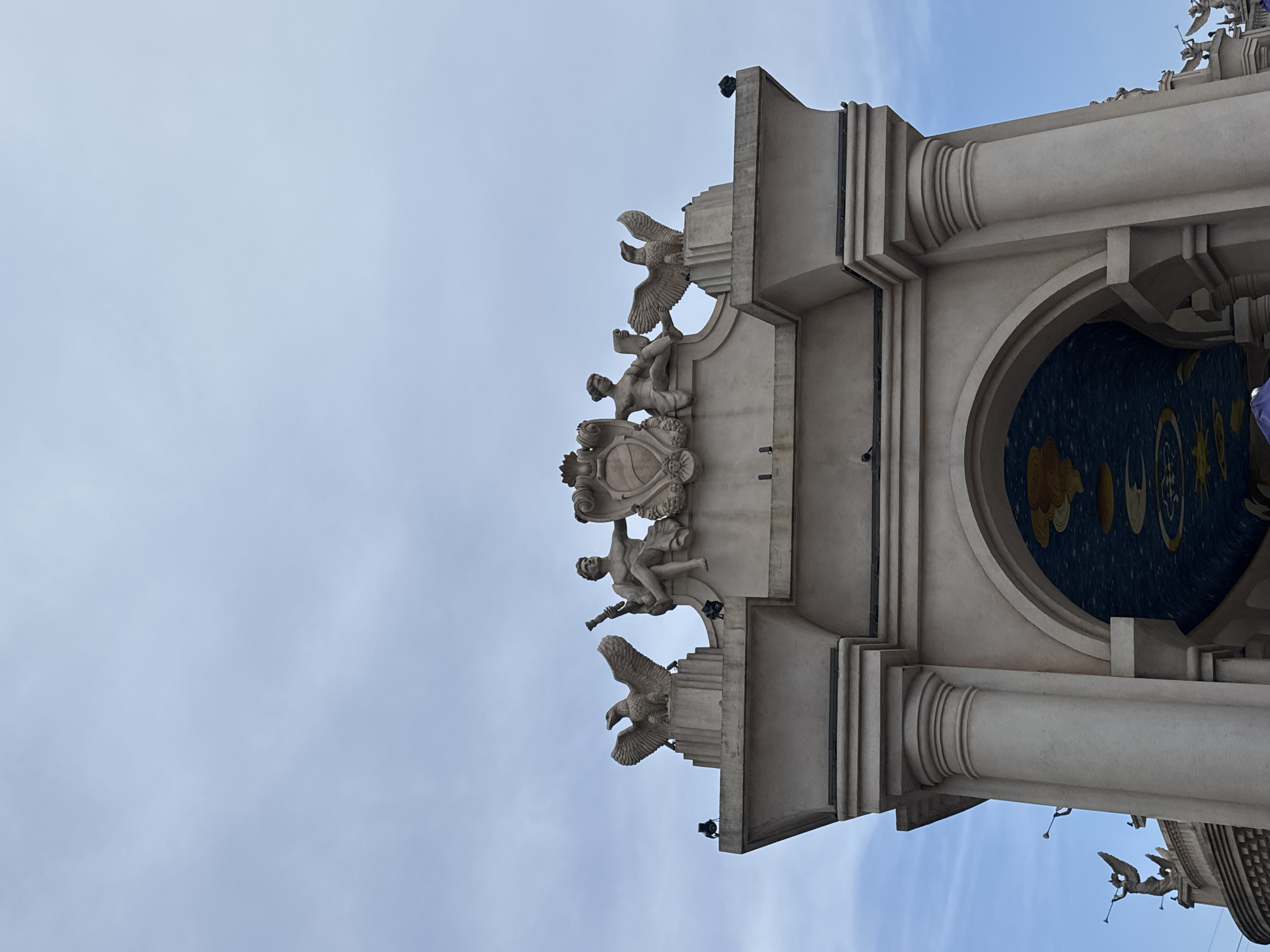 An ornate architectural structure, likely part of a building or monument, stands against a bright, slightly cloudy blue sky. The image is taken from a low angle, looking upwards at a section of the edifice.
The structure is made of light-colored stone or concrete, featuring classical design elements. Prominently displayed is a central heraldic crest or shield, surmounted by what appears to be a crown. This crest is flanked and supported by intricately carved sculptures of human figures, possibly cherubs or putti. Below the crest on each side, large birds, possibly eagles, with spread wings are depicted, adding to the grandeur.
To the right, a large arched opening reveals an interior ceiling or dome decorated with a dark blue background adorned with yellow and gold celestial motifs, including stars, crescent moons, and swirling clouds or suns. The archway is supported by robust columns with fluted details and capitals. Further to the lower right, additional smaller sculptural figures, possibly angels blowing trumpets, can be seen along the roofline.
The daylight is clear and bright, indicating daytime under good weather conditions. The overall aesthetic is one of classical elegance and intricate craftsmanship, characteristic of grand architectural works, often found in themed environments or significant public structures. Given the context, this structure is located in Da Nang, Vietnam. No discernible text is visible in the image.