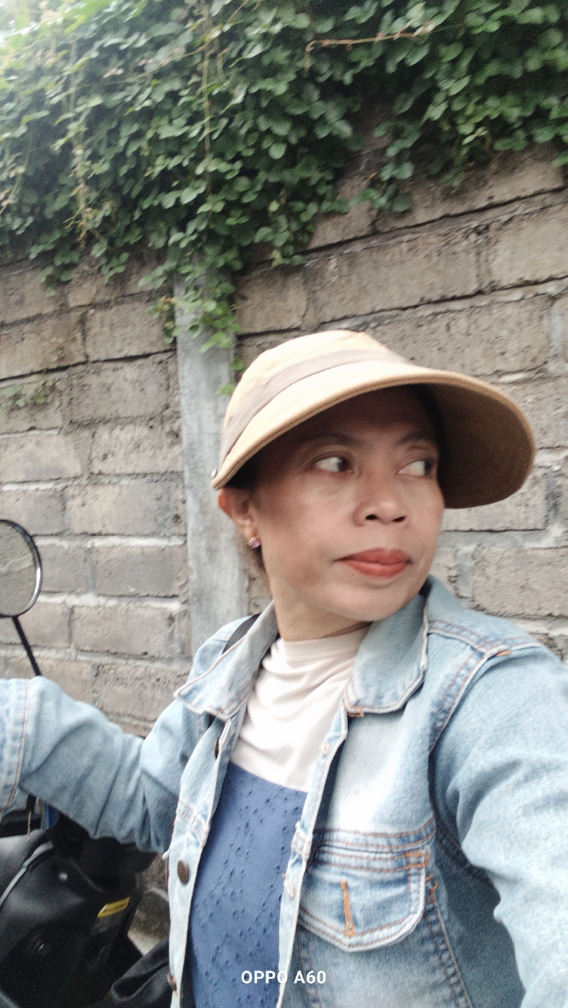 A woman wearing a light brown hat and a light blue denim jacket is photographed from the chest up, looking to her right. She is positioned in front of a brick wall covered in green ivy, with a concrete pillar visible to her left. The woman is also wearing a light beige shirt under her jacket and a blue patterned dress. A black motorcycle is partially visible on the left side of the image. The bottom right corner of the image displays the text "OPPO A60". The scene appears to be outdoors, possibly in a residential area.