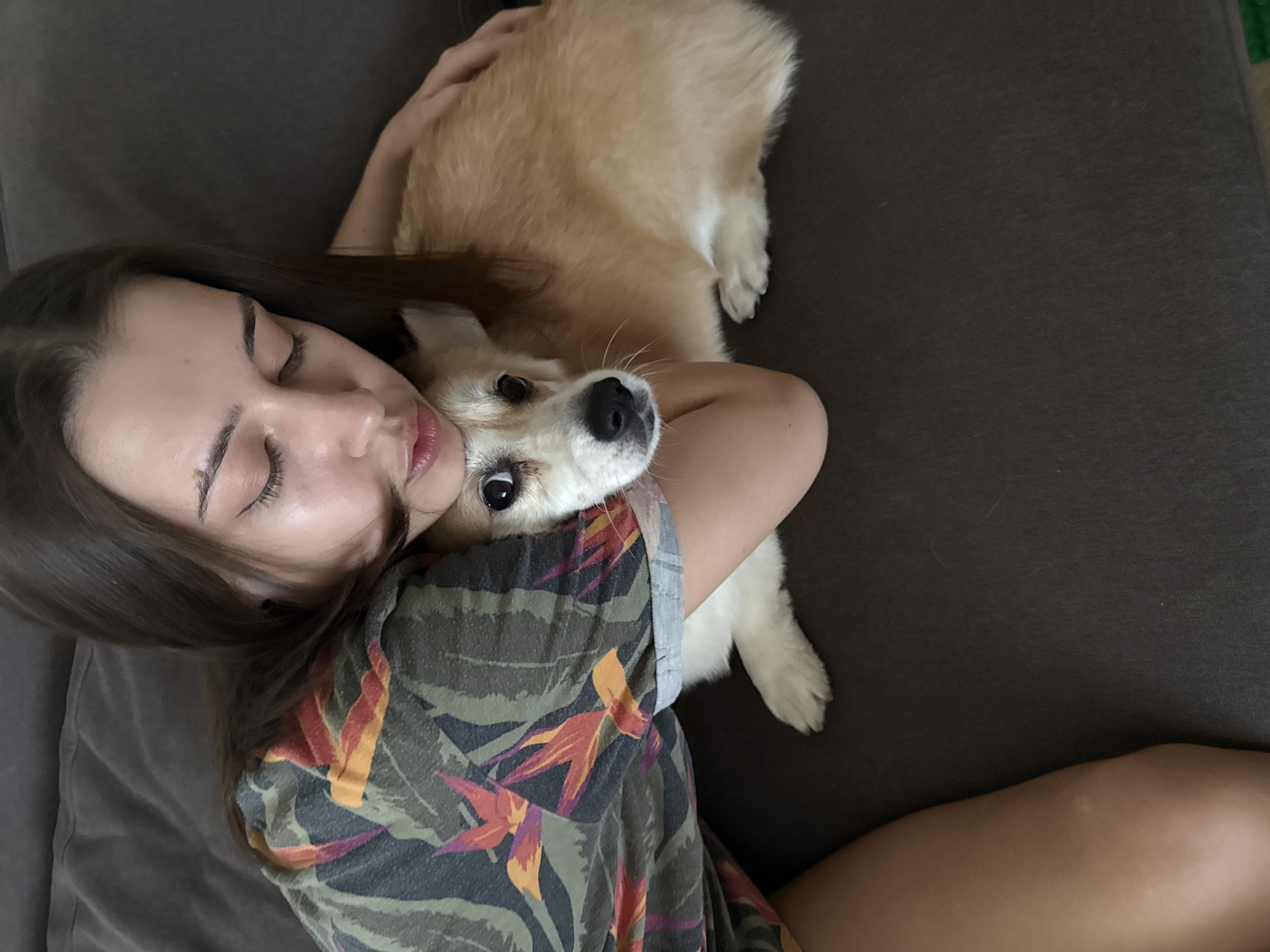 A young woman and a light-colored dog are captured in a close embrace, lying together on what appears to be a dark gray or brown sofa or bed, indoors. The woman, with long dark hair, is positioned on her side with her eyes closed, suggesting she is resting or asleep. She wears a dark green short-sleeved shirt patterned with vibrant orange, red, and purple tropical leaves and flowers. Her left arm is wrapped around the dog.
The dog, which resembles a fluffy corgi with light brown and white fur, is nestled against the woman. Its head rests on her shoulder/arm, and it looks directly at the camera with wide, alert, dark eyes and a black nose. The scene conveys a sense of warmth, affection, and peaceful companionship. The lighting is soft, consistent with an indoor setting, but no specific time of day or weather conditions are evident. There is no visible text in the image. This intimate moment takes place in Krasnoyarsk, Russia.