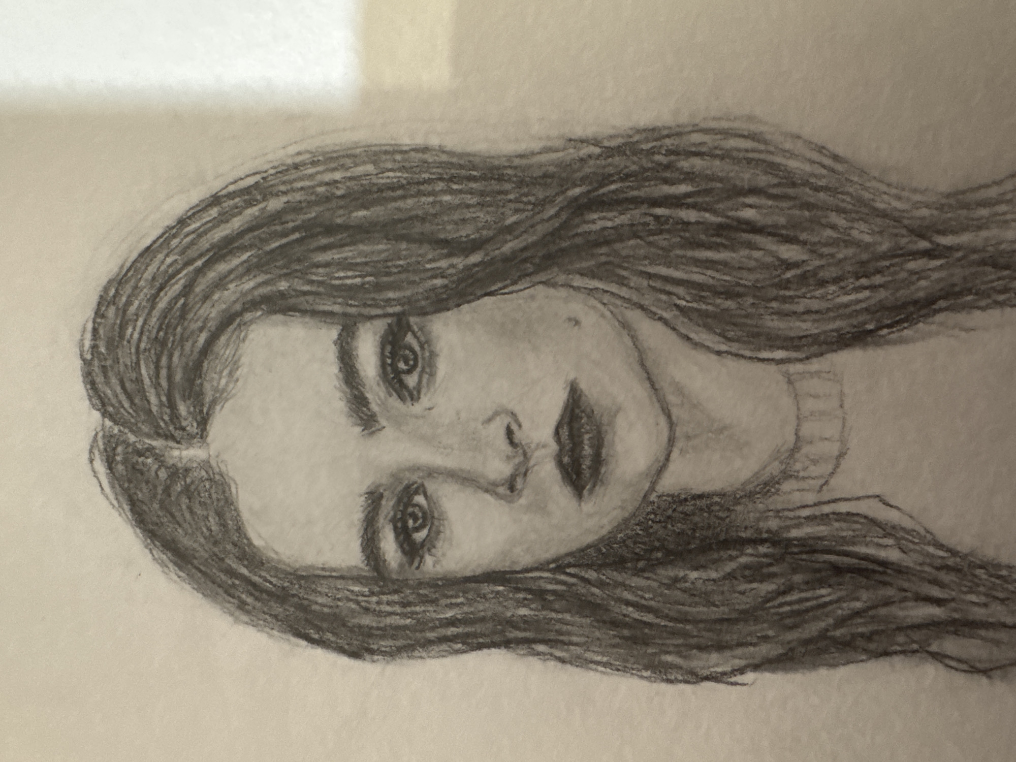 A black and white sketch depicts a young woman's face in a close-up portrait. She has long, dark, wavy hair that frames her face. Her eyes are dark and appear to be looking directly at the viewer. Her nose is slightly upturned, and her lips are full and painted a dark color. There is a small mole on her left cheek. She is wearing a simple sweater with a ribbed neckline. The background is plain and light-colored, with some subtle shading. There is no indication of location in Mikhaylovsk, Russia, or any urban elements in the sketch.
