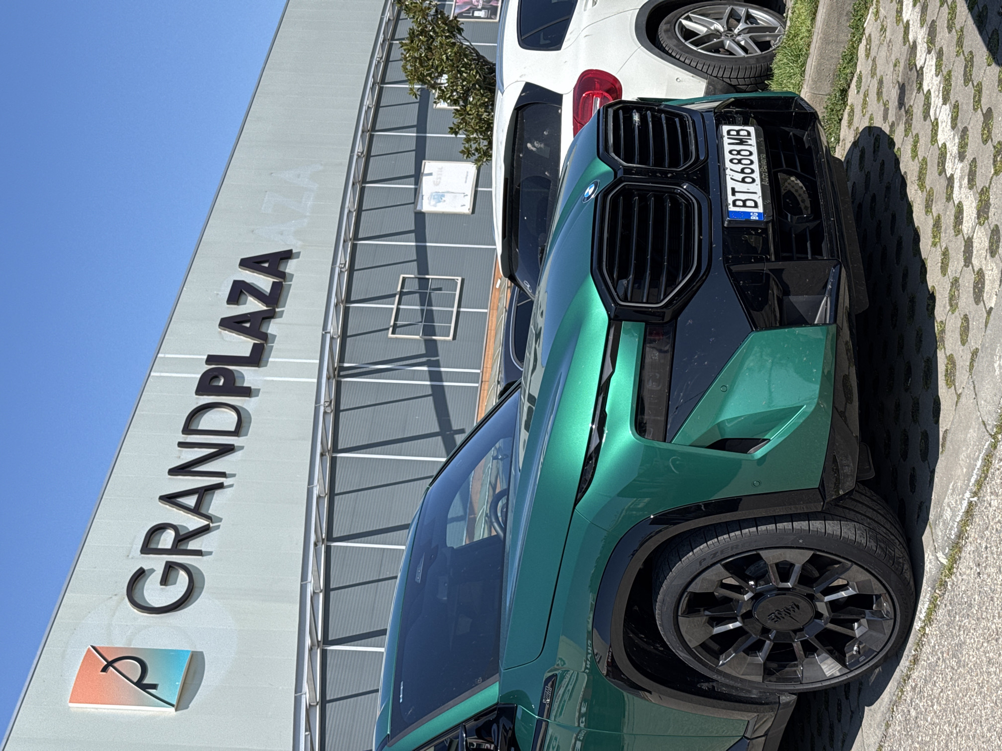 The image captures a green BMW XM parked outside Grand Plaza in Burgas, Bulgaria. The building's "GRANDPLAZA" sign is prominently displayed. A white SUV is parked behind the BMW. The car's license plate reads "BT 6688 MB." The environment is sunny.