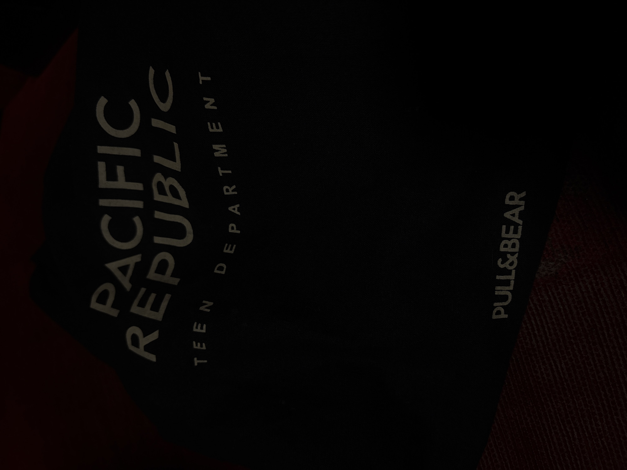 The image shows a dark-colored tag with light-colored text. The text reads "PACIFIC REPUBLIC TEEN DEPARTMENT" and "PULL&BEAR." The tag appears to be associated with clothing. The overall setting and specific location within Kalijaten, Indonesia, are not discernible from the image. There are no people, actions, or interactions visible. The environment is limited to what appears to be the fabric next to the tag.