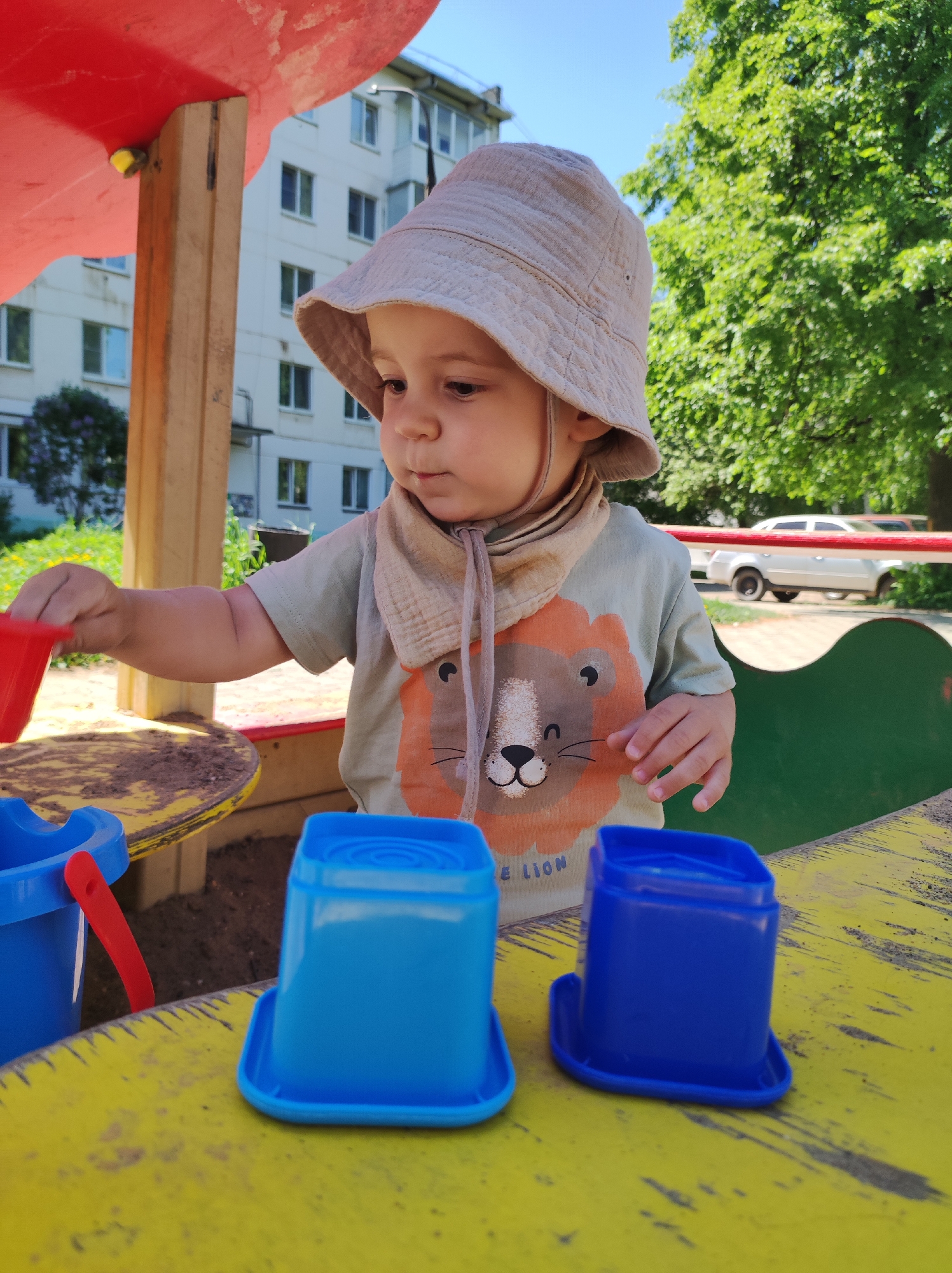A young child, approximately one to two years old, is playing in a sandbox. The child is wearing a beige sun hat and a light green t-shirt with a lion graphic. The child is holding a small red bucket and appears to be filling blue plastic containers with sand. The sandbox is painted yellow and has blue accents. In the background, there is a multi-story building with white walls and numerous windows. Green trees surround the play area. A white vehicle is partially visible parked in the background. This photograph was taken in the city of Chaikovsky, Russia.