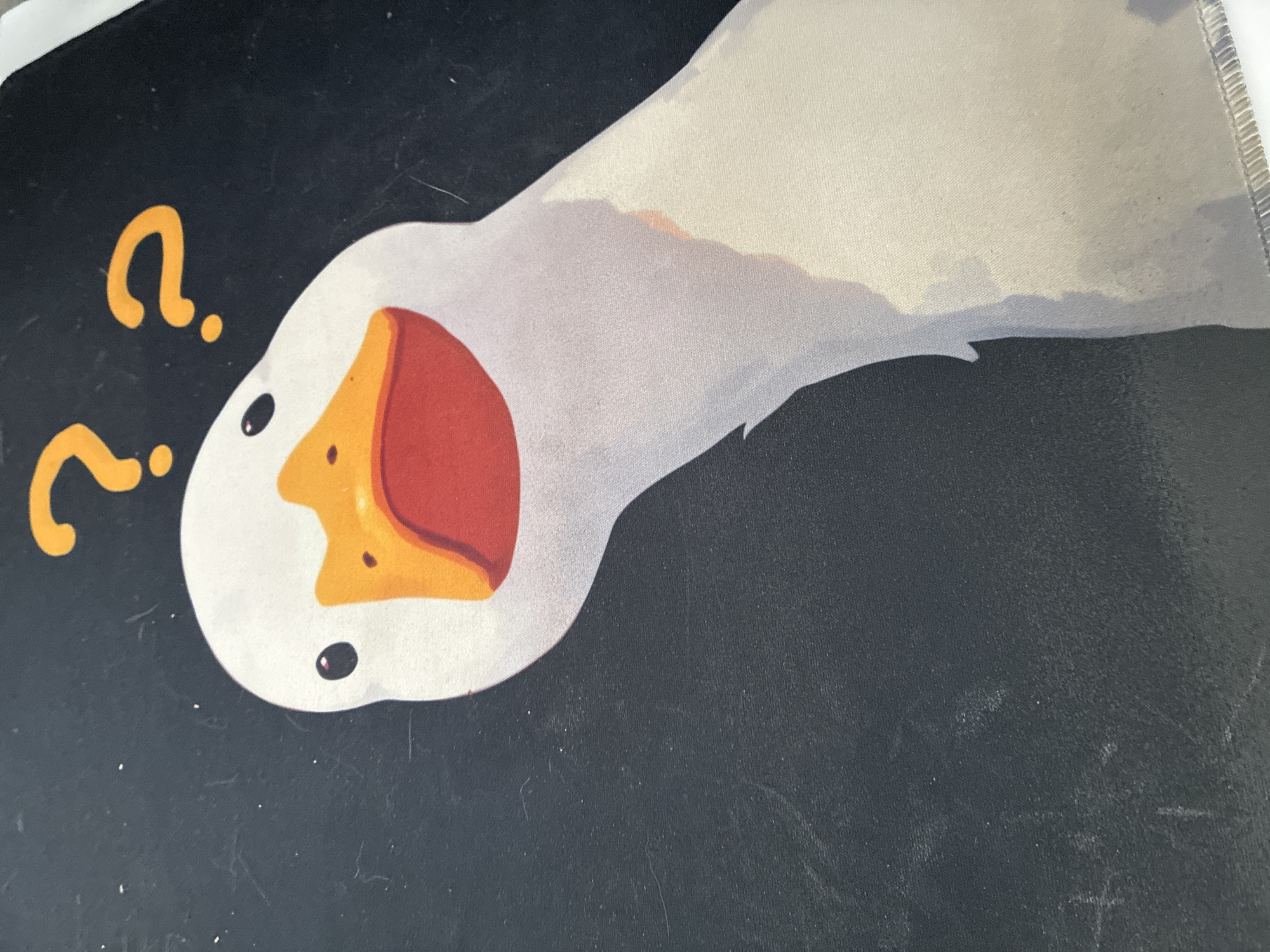 The image displays a close-up view of a white goose with its head tilted and a questioning expression, indicated by two orange question marks above its head. The goose has a prominent orange beak and a red mouth. The background is black, and the image appears to be printed on fabric, possibly a mousepad, given the slight sheen and visible texture. There is no indication of the image's location in Kyiv, Ukraine, or any people, actions, or interactions. The scene is a static graphic.