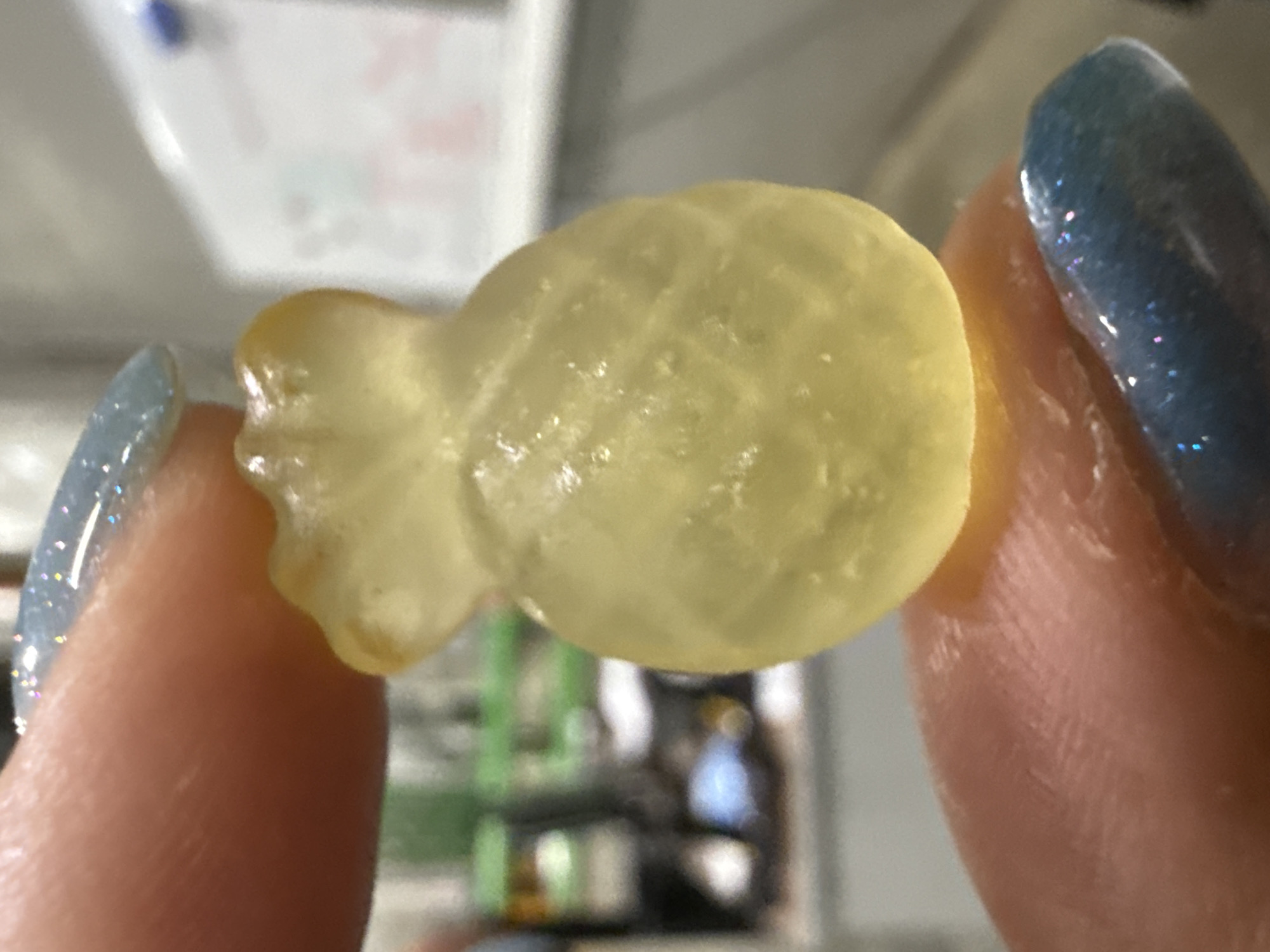 A yellow, fish-shaped gummy candy is held between two fingers. The fingers have light skin and are adorned with painted fingernails. The nail on the left appears to be pale blue and speckled, while the nail on the right is a darker, glittery blue. The background is blurred, but suggests an indoor setting with various objects and containers. The picture does not show or suggest any connection to Lupanovo, Russia.