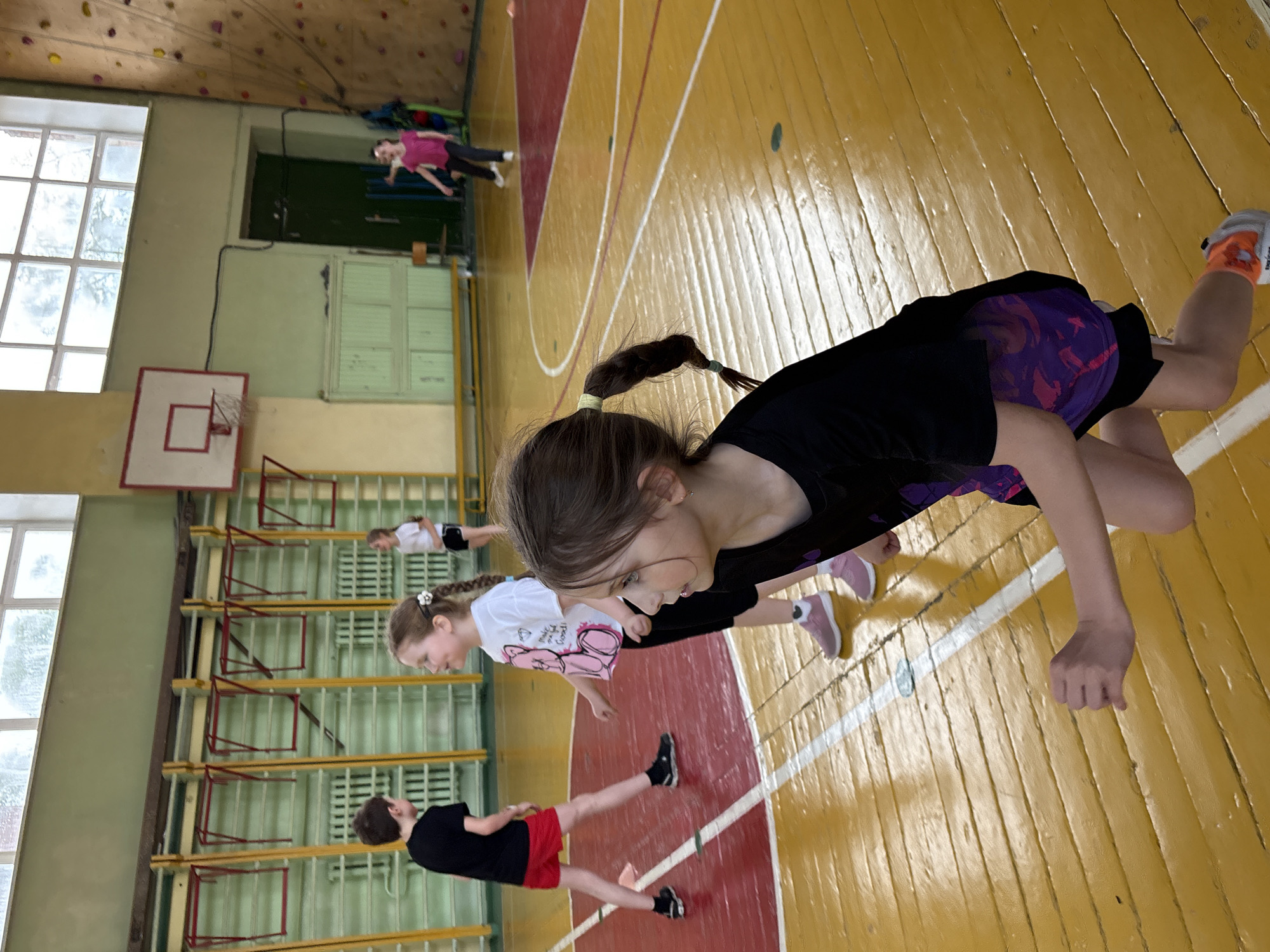 The image shows a group of children exercising in a gymnasium. The gymnasium has wooden floors with white lines, basketball hoops, and a rock climbing wall. There are at least five children present, all in athletic clothing. The gymnasium is located in Izhevsk, Russia.