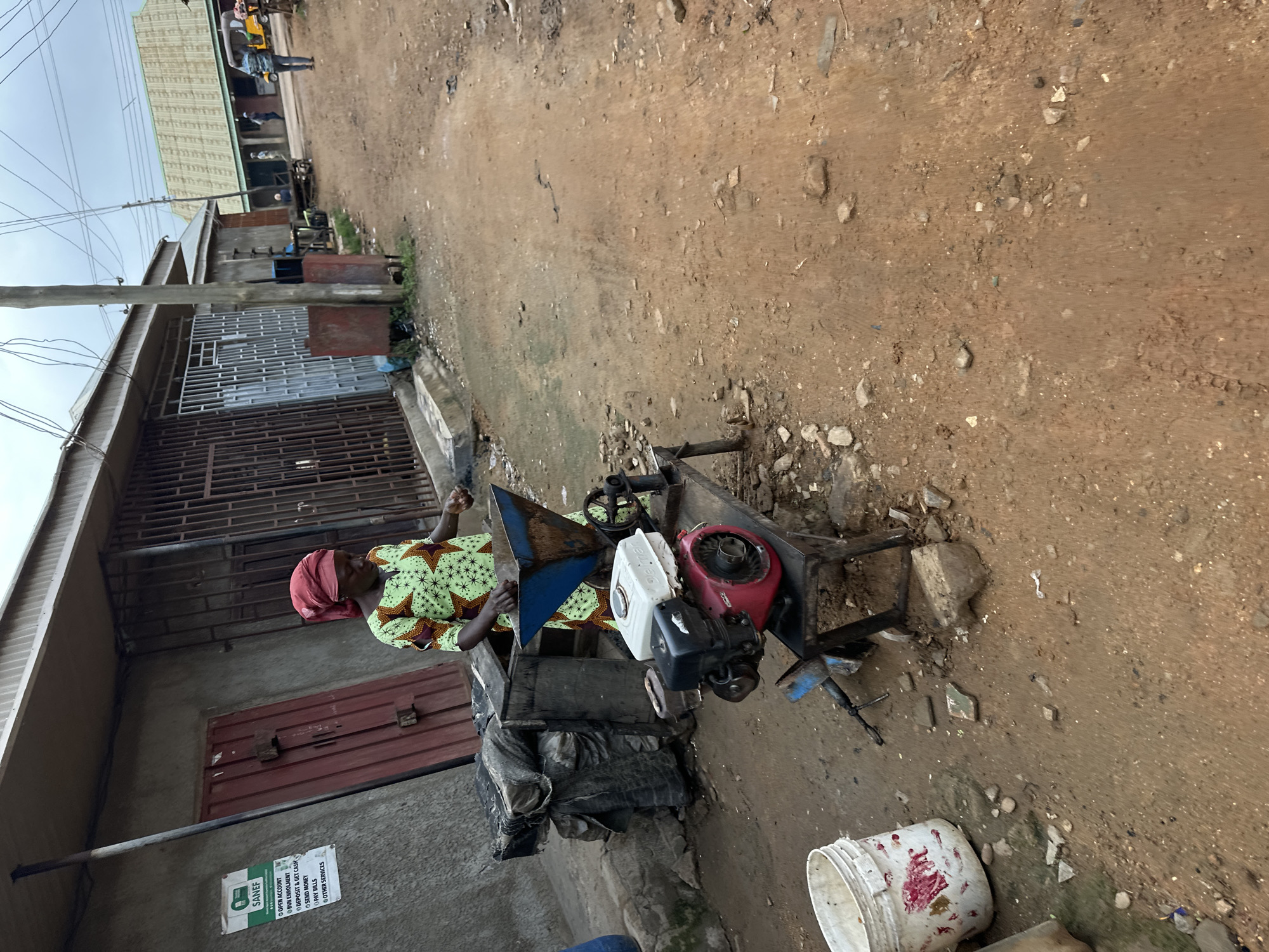 The image depicts a street scene in Narayi, Nigeria. In the foreground, a woman in a patterned green and yellow dress and a red headscarf stands beside a metal grinding machine powered by a red and white engine. The machine has a blue hopper on top. To the left of the woman and machine, a white bucket with red stains sits on the ground. A dark, tarp-covered object is also visible to the left. The ground is dirt and strewn with small rocks. In the background, there are buildings with corrugated metal roofs and barred windows, typical of an urban African setting. A dirt road continues into the distance, with a few people and a motorized tricycle visible. Power lines are strung overhead. A sign on the wall behind the woman lists various services. The overall impression is of a busy, somewhat dusty street in a commercial or residential area.