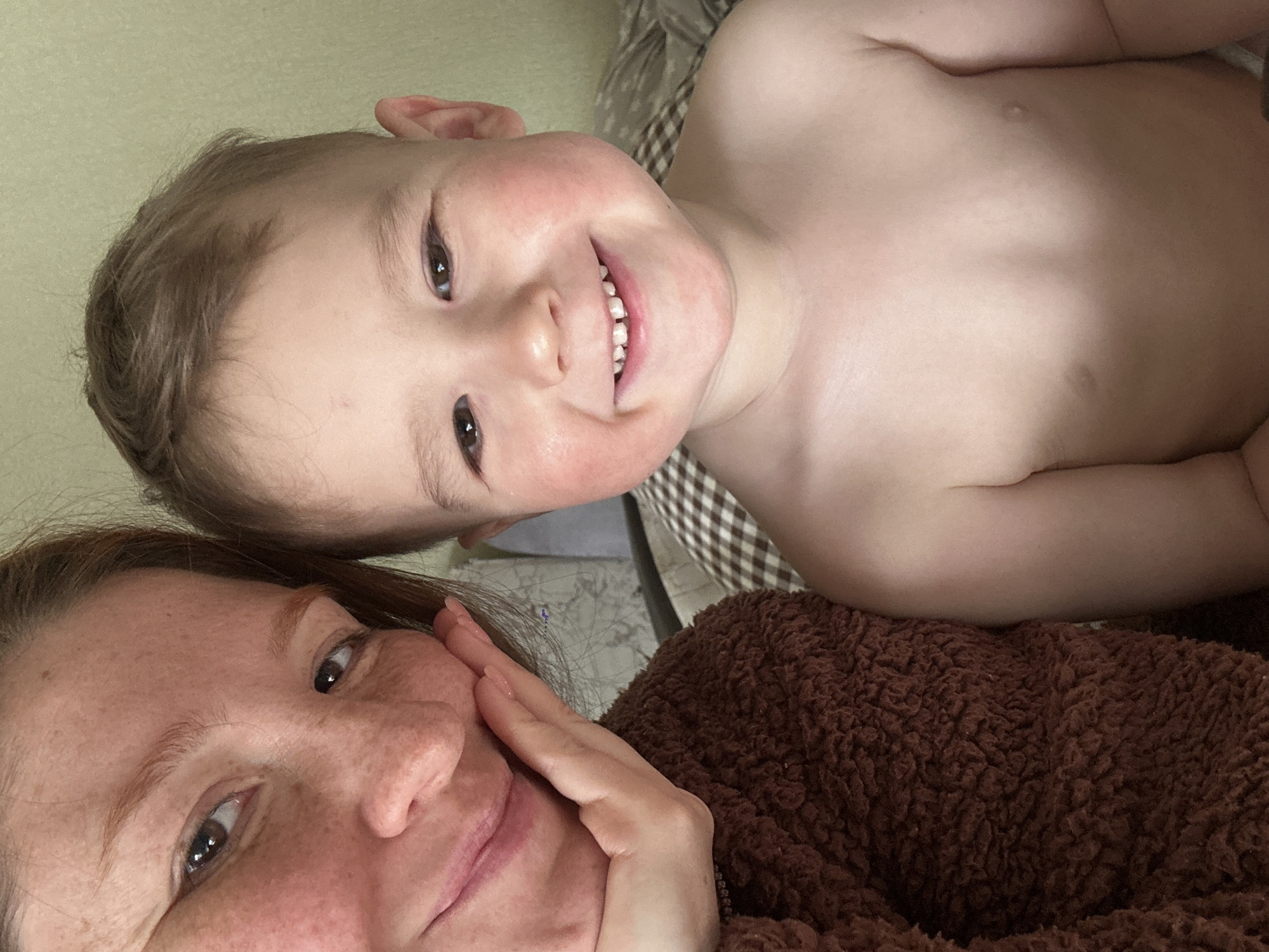 A close-up shot features a woman and a young child. The woman, with fair skin and reddish hair, is wearing a brown fuzzy top and has her hand on her face. The child, positioned above her, is shirtless and has fair skin and light brown hair. Both are smiling. The background is blurred, but a plaid pattern suggests bedding or a blanket. There is no contextual information to suggest location, and it is not possible to identify the location of the photograph as Nizhny Novgorod, Russia.