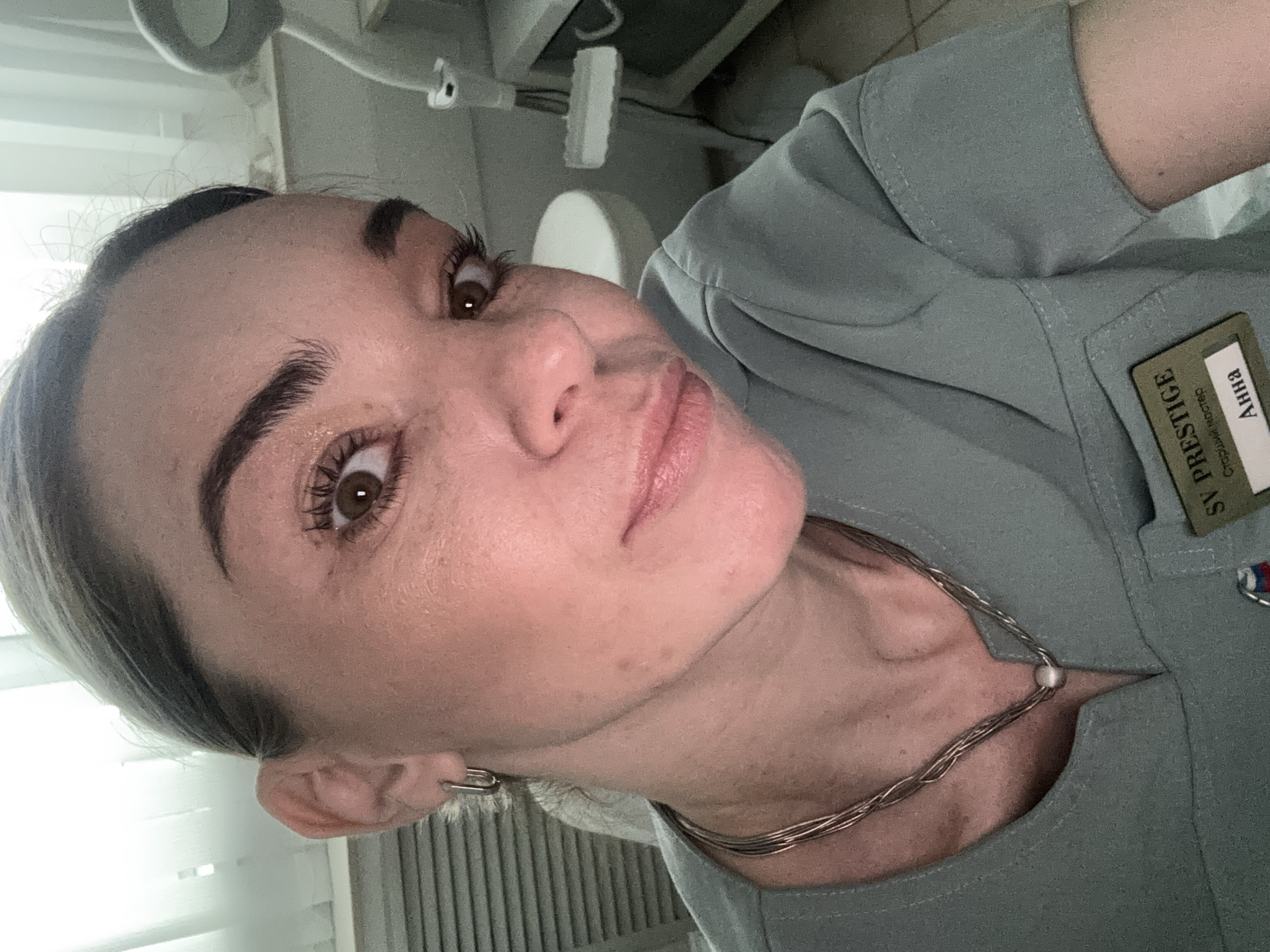 A close-up selfie shows a woman wearing a light green uniform. Her name tag reads "Anna SV PRESTIGE" in Russian. She has light skin, brown eyes, and dark eyebrows. She wears a multi-strand silver necklace and a silver earring is visible. The background suggests a clinical setting with medical equipment and window blinds. Location: Unknown, Unknown.