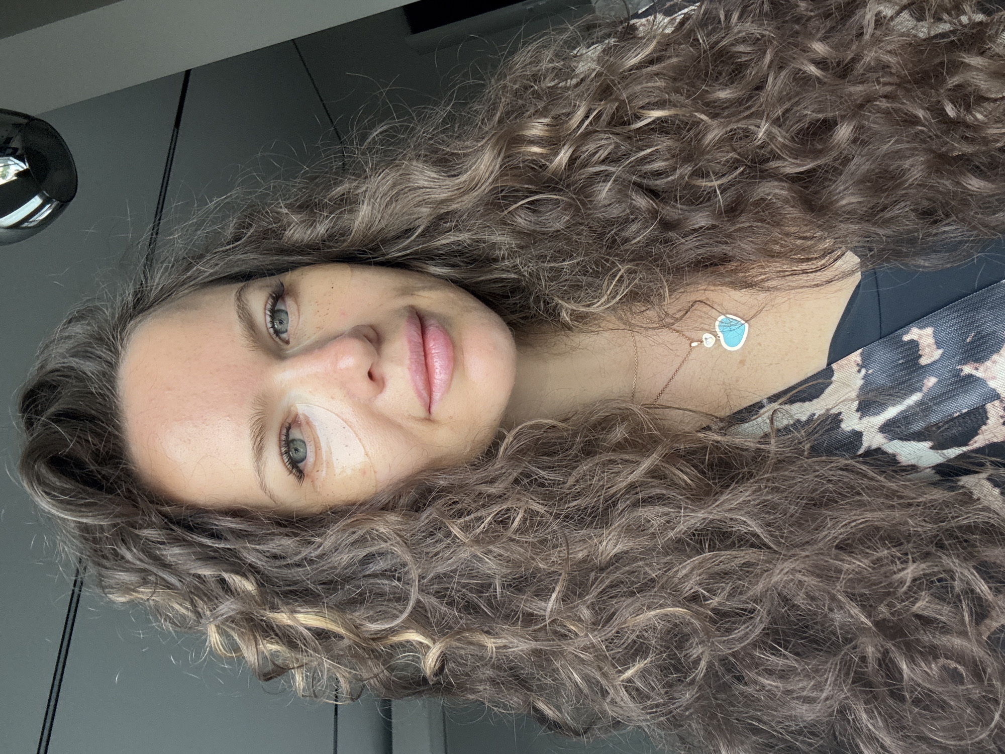 A woman with long, curly brown hair is shown in a close-up selfie. She is wearing eye patches and a necklace with a blue heart pendant. Her clothing has a leopard print pattern. The background appears to be an indoor setting with a ceiling light fixture visible. The image was taken in Dolgoprudny, Russia.