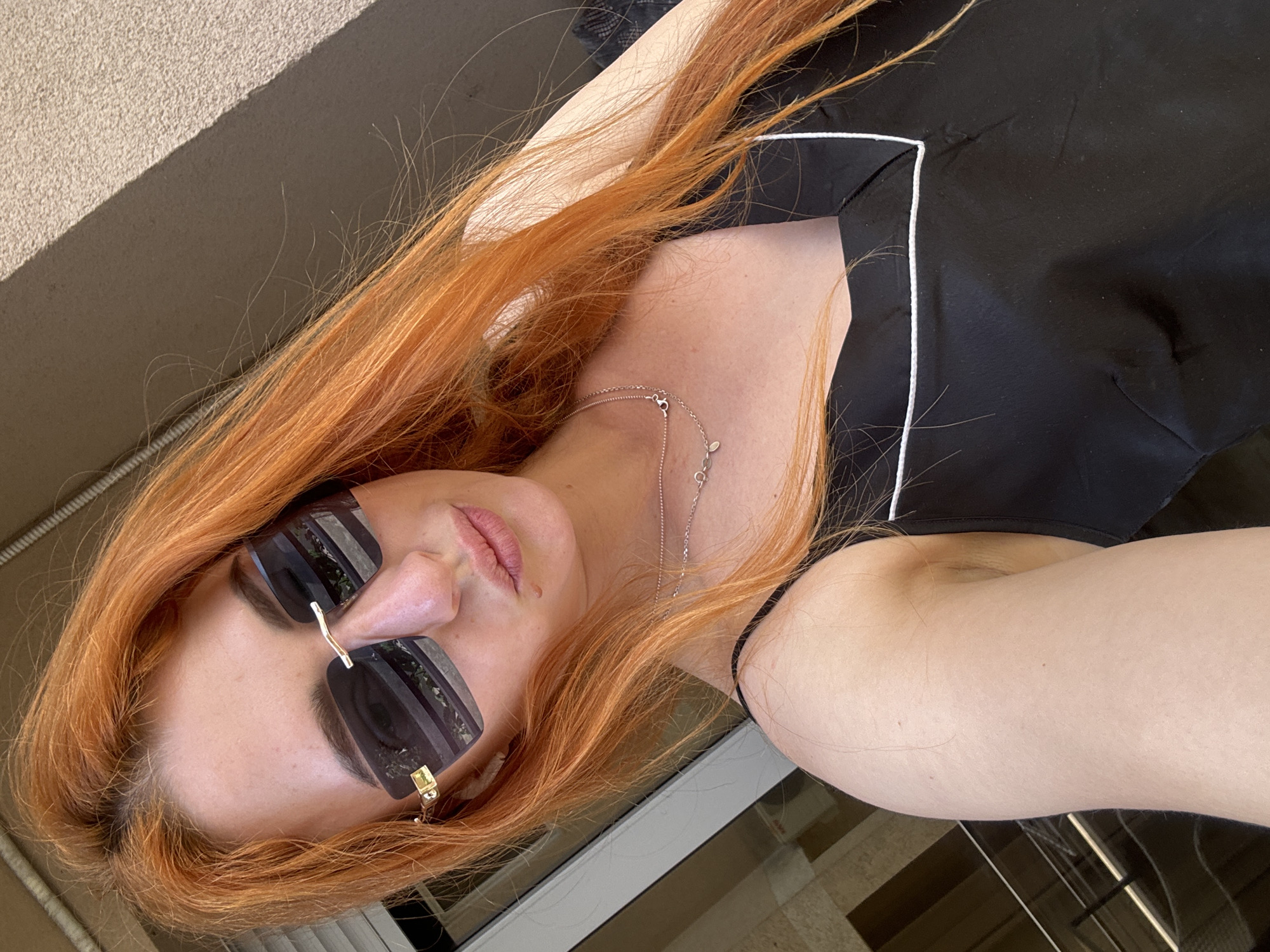A woman with long, red hair, wearing sunglasses and a black top with white trim, is positioned at an angle in an indoor location that appears to be a building in Budva, Montenegro. She is also wearing a silver necklace. The background includes a wall and flooring that show a modern or simple architectural style. The angle of the image indicates it might be a selfie.