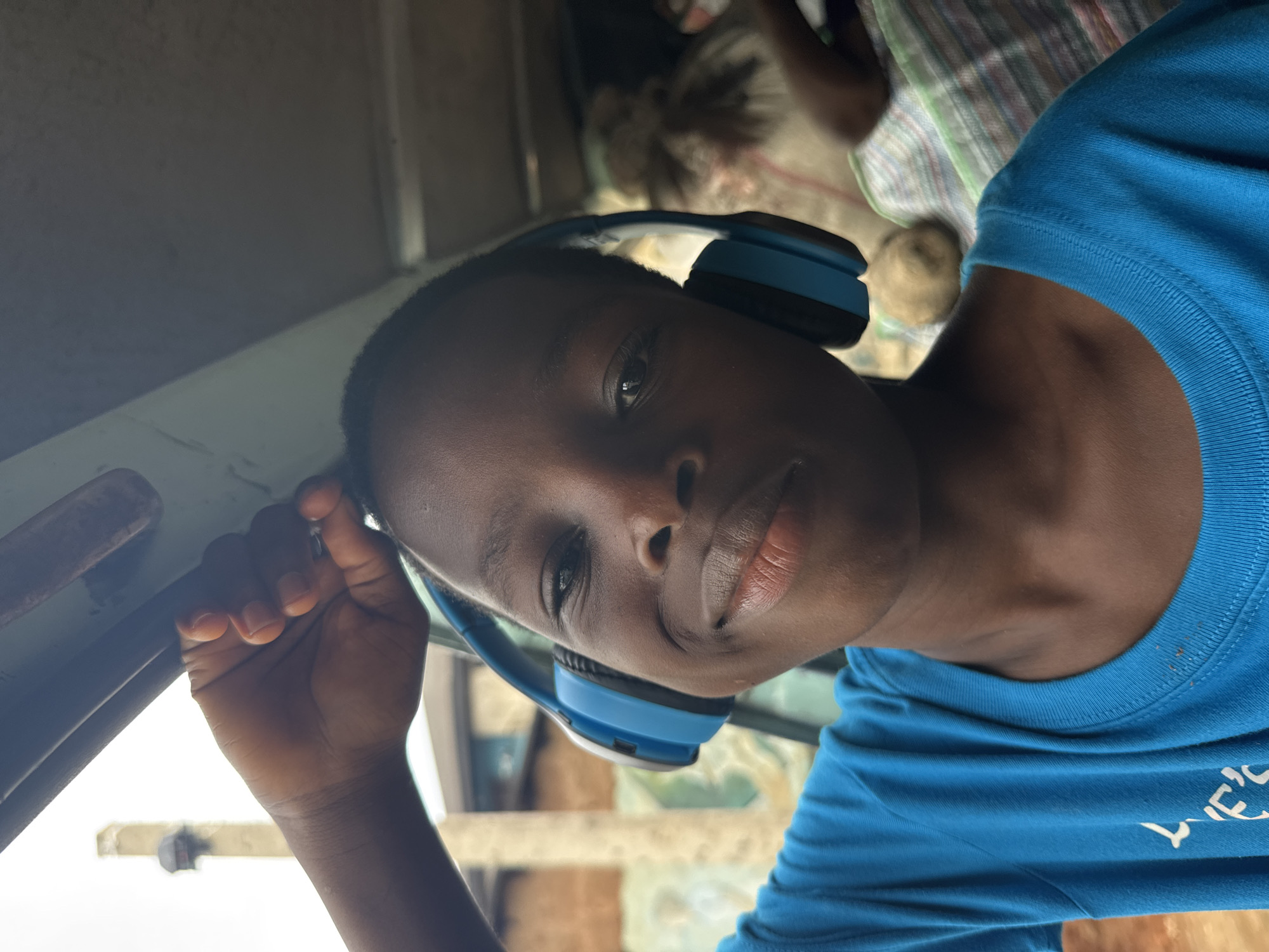 A person is centered, wearing a blue t-shirt and blue headphones. The person is holding onto a silver bar, likely a grab bar. They are inside a vehicle, looking at the camera. There is another person with plaid clothing in the background. Based on the style of the vehicle and the surroundings, the location is likely in Onitsha, Nigeria.