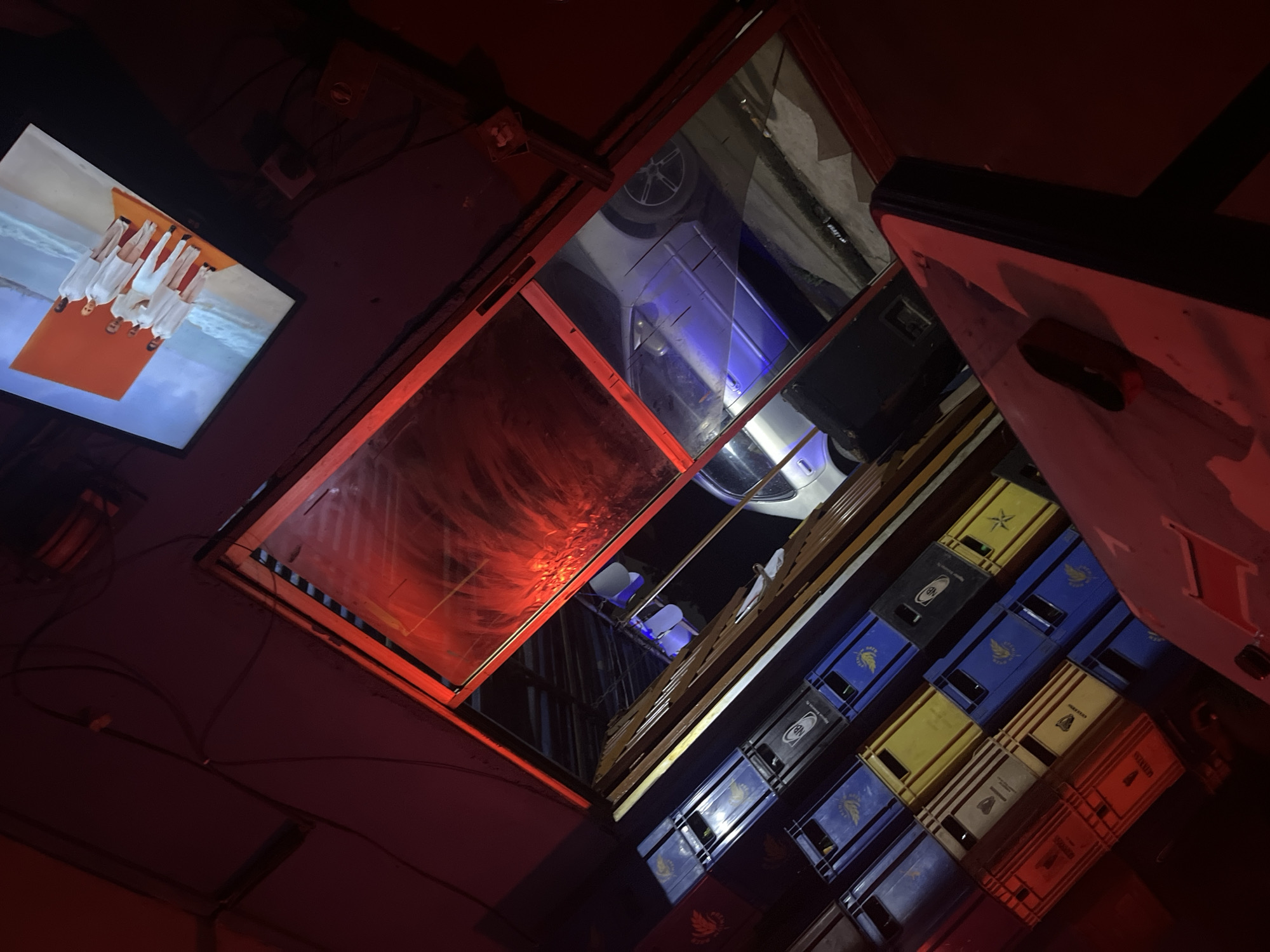 The image appears to be taken inside a dimly lit establishment, possibly a bar or a storage area, with heavy red lighting casting an atmospheric glow. In the background, a television screen displays an image of five individuals wearing white clothing standing on an orange platform against a backdrop of water and sand.
The foreground and middle ground are dominated by stacks of plastic crates, some colored blue, yellow, and red, suggesting they might be used for storing beverages. Through a glass partition, a silver car is visible parked outside, illuminated by what appears to be blue lighting. The overall scene is somewhat cluttered, with visible wires and what looks like a speaker box. The presence of the crates and the overall ambiance suggest a commercial setting.
Given the specific request, it is impossible to determine the location in the city of Jos, Nigeria, from the visual information provided in the image alone. There are no identifiable landmarks, signs, or contextual clues that would link this scene to a particular place within Jos.