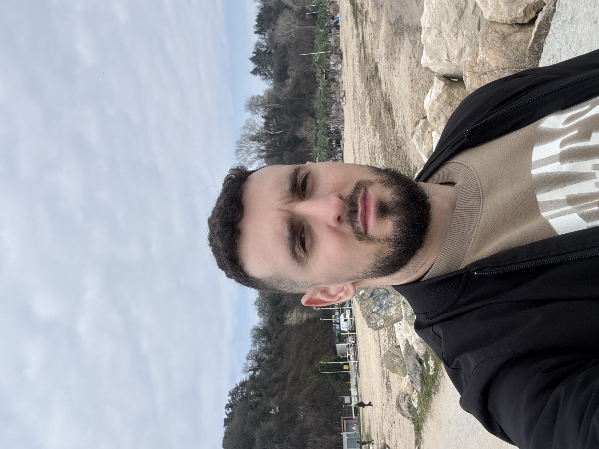 A man with dark hair and a beard is taking a selfie. He is wearing a beige sweater with a white pattern and a black jacket. The background suggests he is at a beach or near the coast. Trees are visible on a hillside in the background. Based on the specific terrain and hillside vegetation, the location is likely in the city of Varna, Bulgaria. The sky is overcast. There are people walking on the beach behind him. There are also sun umbrellas visible in the background.