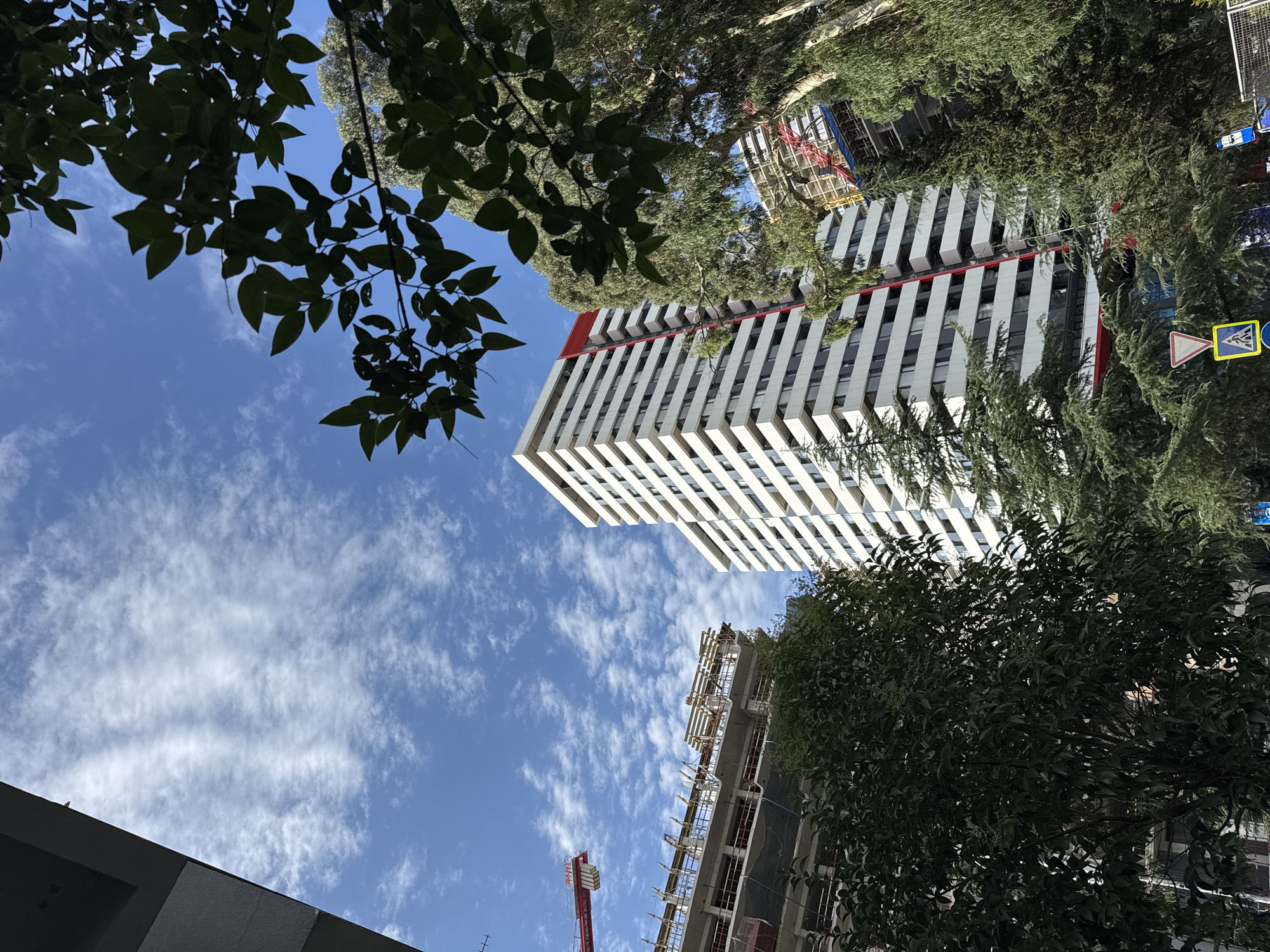 In Batumi, Georgia, this low-angle shot captures a clear blue sky with scattered cirrus clouds above a mix of urban elements. To the left, the edge of a modern building is visible. In the center, a tall, white residential building with horizontal dark windows and a red accent strip dominates the view. In front of and to the right of this building, lush green trees with dark leaves and needle-like foliage partially obscure the scene. Further left, a building under construction is seen with scaffolding and a red crane extending into the sky. Street signs, including a triangular warning sign and a rectangular yellow sign with a blue symbol, are visible at the bottom of the frame.