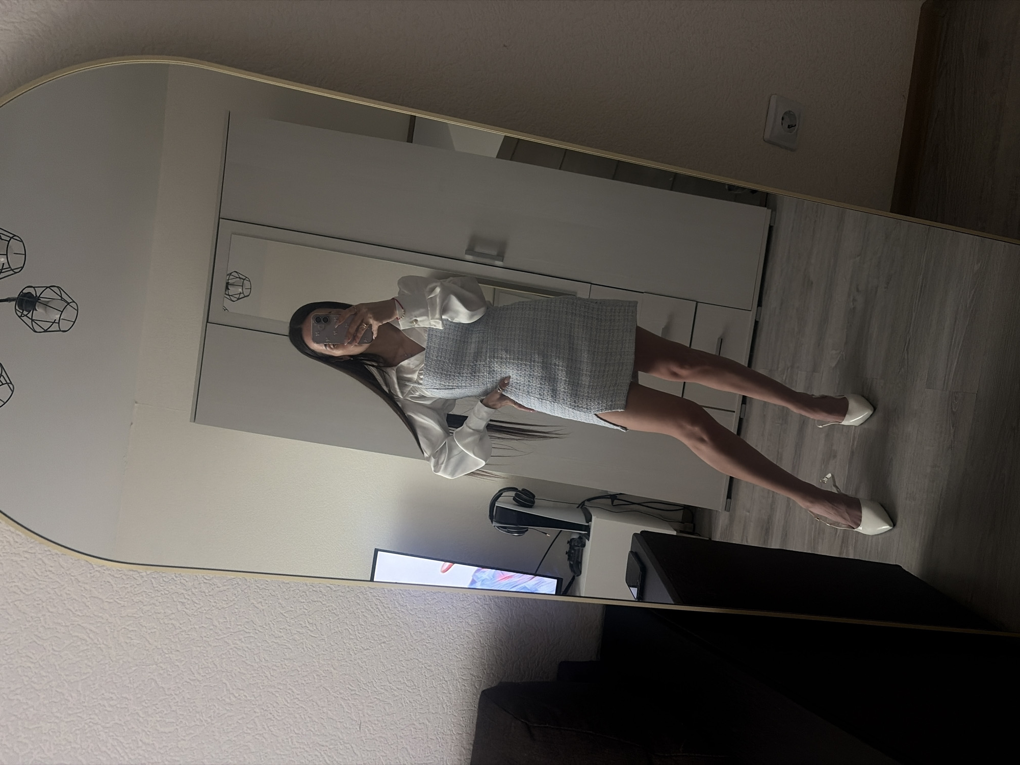 A person is captured in a mirror selfie, standing sideways to the camera, showcasing their full outfit. The individual has long dark hair and is holding a smartphone with a light-colored, decorated case up to their face to take the picture. They are wearing a white blouse with prominent, voluminous cuffed sleeves and a light blue or grey textured mini-dress or skirt. White, pointed-toe high heels with an ankle strap adorn their feet. Rings are visible on their right hand.
The large, arched-top mirror with a gold-toned frame reflects a modern indoor space, likely an apartment in Saint Petersburg, Russia. The walls are light-colored, and the floor features light grey wood-look laminate or planks. Reflected objects include a white closet or wardrobe with silver handles, a black metal cage-style pendant light fixture with multiple exposed bulbs, and a white European-style electrical outlet on the wall. In the background of the reflection, a television screen displays a colorful abstract image, and a white PlayStation 5 console with a black headset rests on a surface near it. Part of a dark-colored sofa or piece of furniture is visible in the lower foreground of the reflection. The lighting suggests an indoor setting, likely illuminated by artificial light. The entire image is oriented 90 degrees clockwise, so the person appears to be standing horizontally.