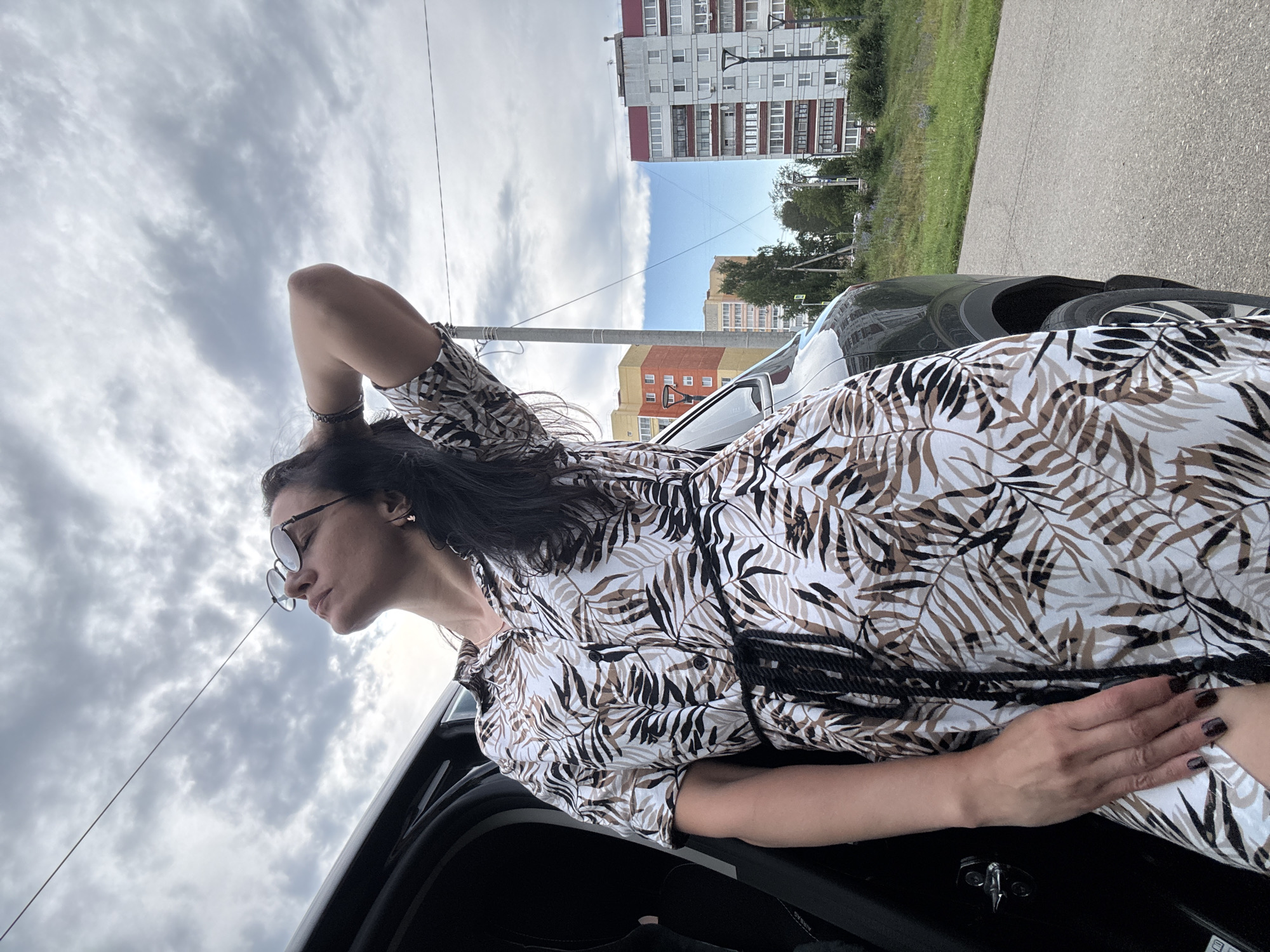 A woman with dark hair and sunglasses leans against an open car door, her right arm resting on her stomach and her left arm behind her head. She wears a white dress with a brown and black leaf-like pattern and dark nail polish. The scene appears to be outdoors in an urban environment. In the background, several multi-story buildings are visible, suggesting a residential area. A paved road and a grassy area with trees are also present. The sky is cloudy, indicating overcast weather. The image is oriented vertically, with the top of the frame pointing upwards towards the sky. No visible text is present in the image.