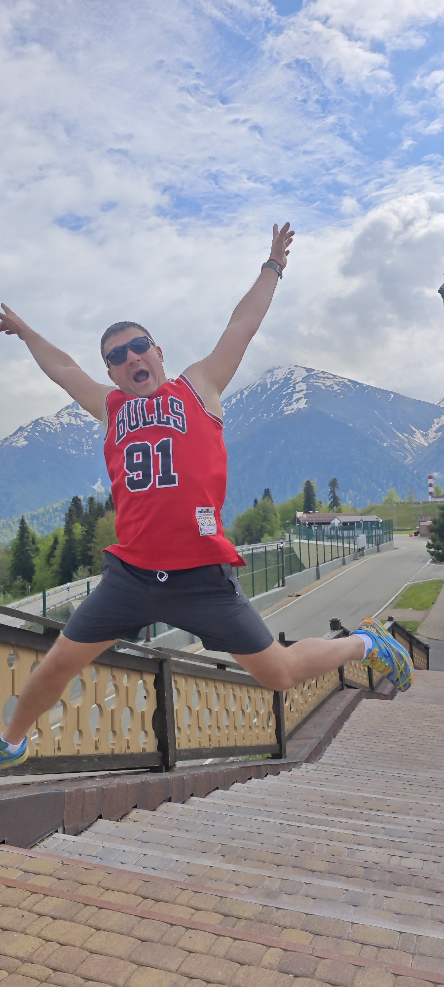 A man is suspended mid-air in a dynamic pose, wearing a red Chicago Bulls #91 jersey, dark shorts, sunglasses, and brightly colored running shoes. He has his arms spread wide and legs extended in a split jump, his mouth open in a yell. He is positioned over a wooden railing and a series of tiled steps. Behind him, a road winds through trees, and snow-capped mountains rise in the background under a partly cloudy sky. The scene is set in the city of Unknown, Russia. A white and green building is also visible in the background.