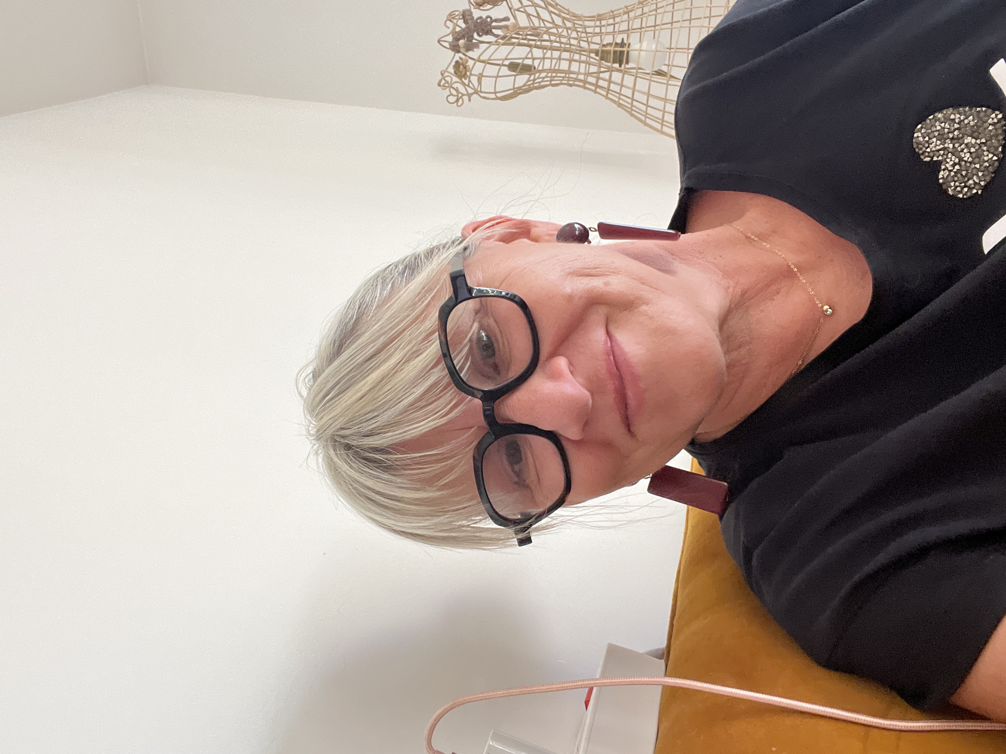 A woman with blonde hair and glasses is looking at the camera. She is wearing a black t-shirt and large earrings. She is sitting on a yellow couch in front of a white wall. A lamp is to her left, and a wire mannequin is to her right. The scene is an indoor setting, likely a living room. There are no discernible elements that place the scene specifically in Montélimar, France.