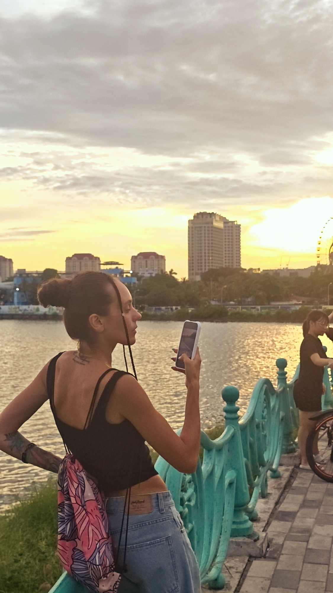 In an urban setting during sunset, a woman with braided hair pulled into a bun stands on a paved walkway next to a body of water. She wears a black crop top, blue jeans, and a colorful drawstring bag. In her right hand, she holds a smartphone, seemingly taking a picture. The backdrop features a city skyline with tall buildings and lush greenery. To the right, another woman in a dark dress is visible, partially obscured by a bicycle. The scene suggests a recreational area, possibly a park or a waterfront promenade, within Hanoi, Vietnam, given the architectural styles and natural elements. The sky is painted with warm hues of orange and yellow, indicating the end of the day.