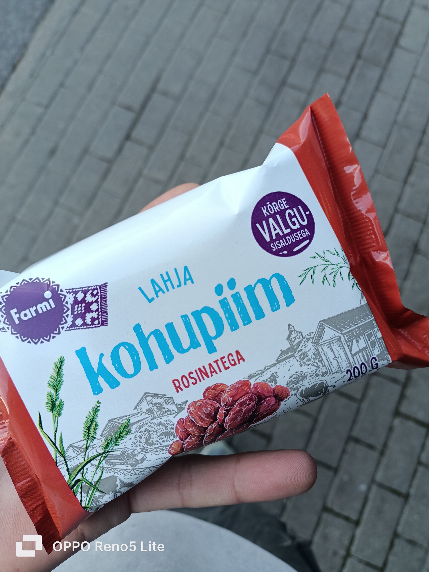 The image shows a close-up of a food package held by a person's hand. The package is red and white with blue and purple text. The text on the package includes "Farmi", "LAHJAKOHUPIIM", "KÕRGE VALGU SISALDUSEGA", "ROSINATEGA", and "200 G". There are illustrations of wheat, farm buildings, and cows on the package. The background is a grey paved surface. The bottom right corner of the image has a watermark that reads "OPPO Reno5 Lite". The scene is outdoors on a paved surface. The image is taken from a slightly elevated angle, looking down at the package. There is no specific indication of the location being in Tallinn, Estonia.