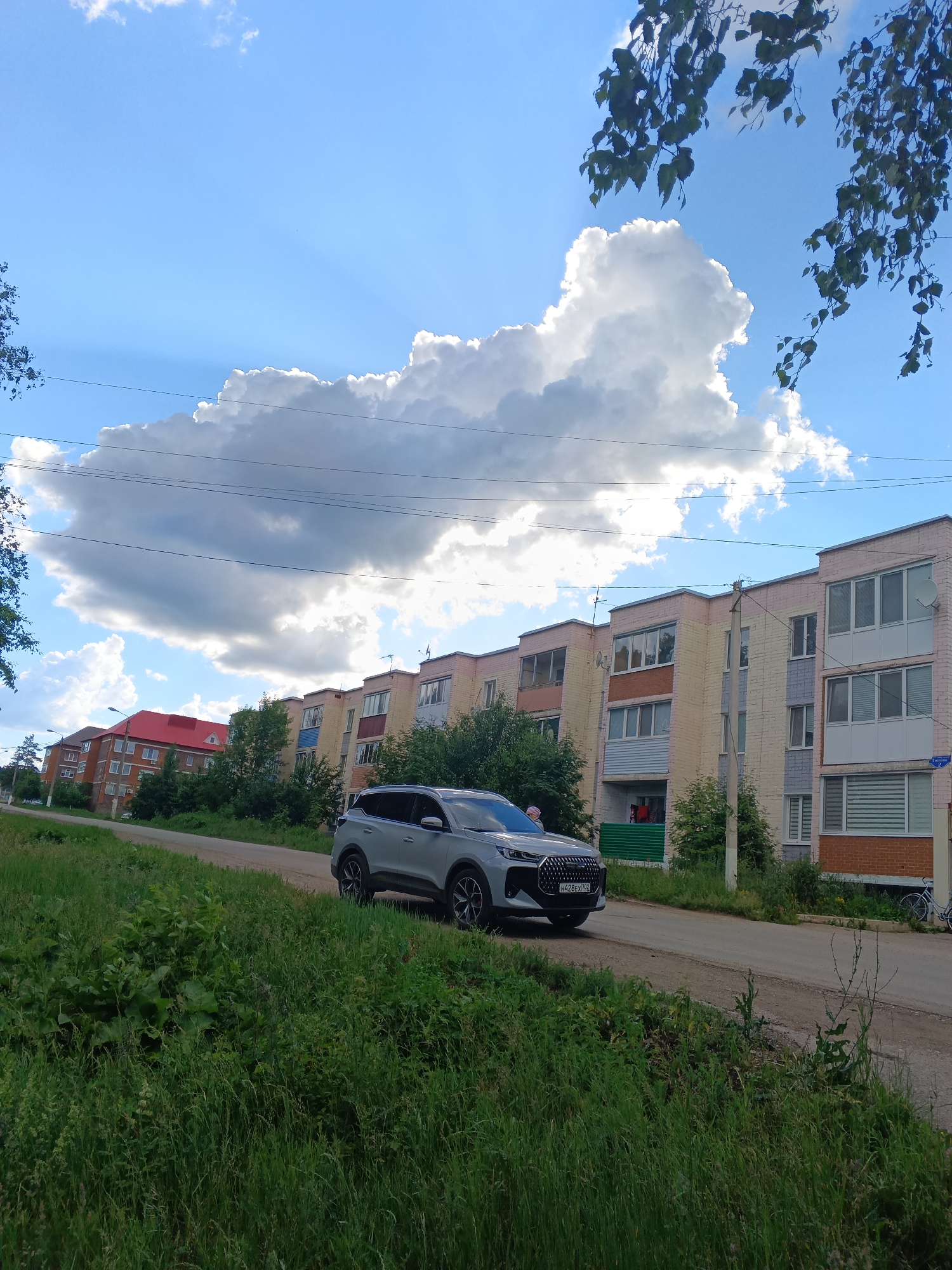 In Tolbazy, Russia, a silver SUV is parked on a dirt road next to a grassy embankment. The SUV faces right, partially obscuring a row of apartment buildings. The buildings are multi-story structures with light-colored facades, some featuring balconies. Green trees are interspersed between the buildings. A large, fluffy white cloud dominates the blue sky. The foreground is filled with lush green grass and some taller weeds. In the background, more apartment buildings and trees are visible, suggesting a residential area within the town.