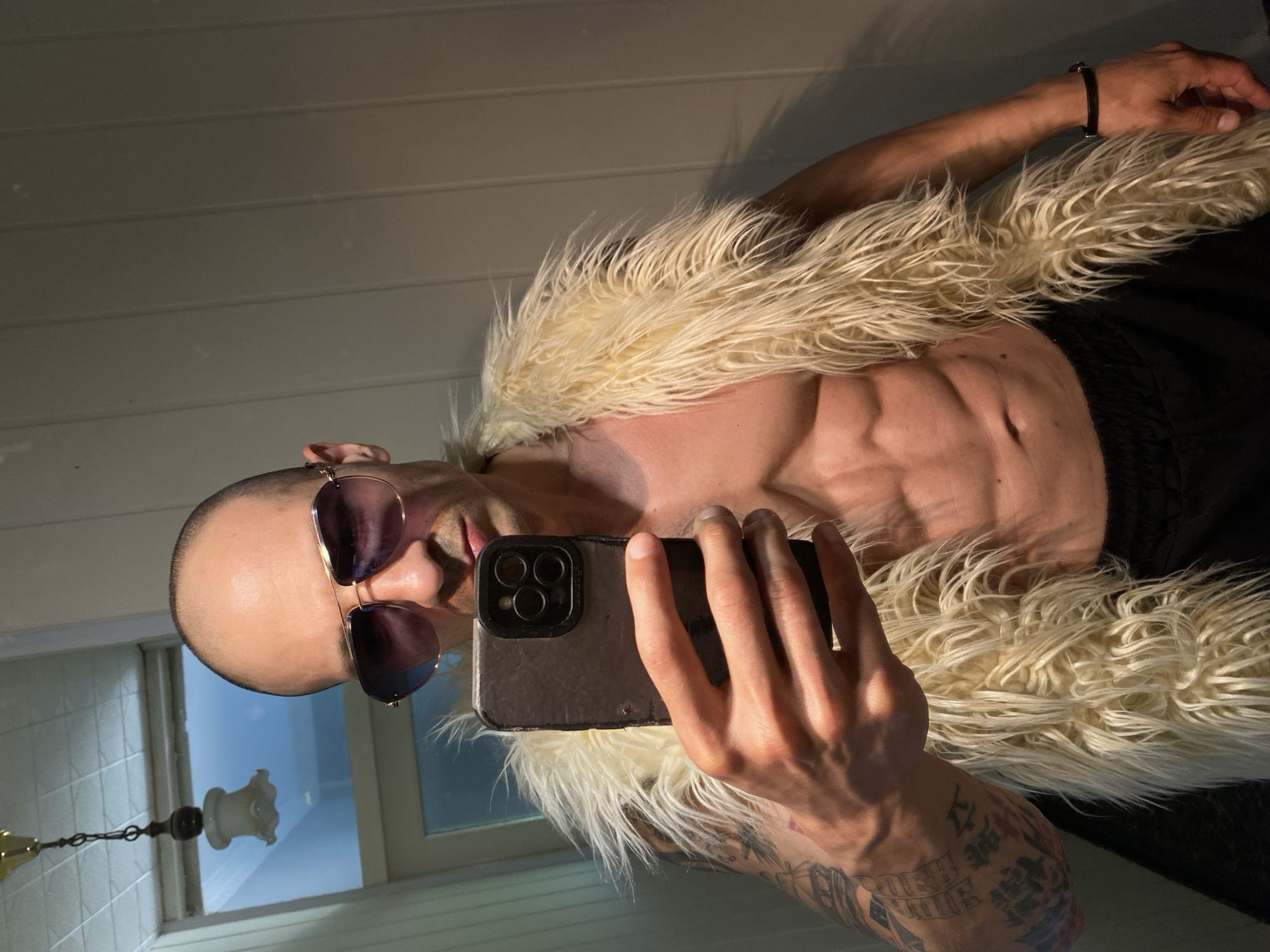 A bald man with sunglasses is taking a selfie in what appears to be a bathroom. He's shirtless, wearing a light-colored, shaggy fur vest. Visible tattoos are on his left arm. He is holding a smartphone with three visible camera lenses. The background includes white paneling, a window reflecting a chandelier, and a tiled wall. There is no information to suggest that the image was taken in Pleven, Bulgaria.