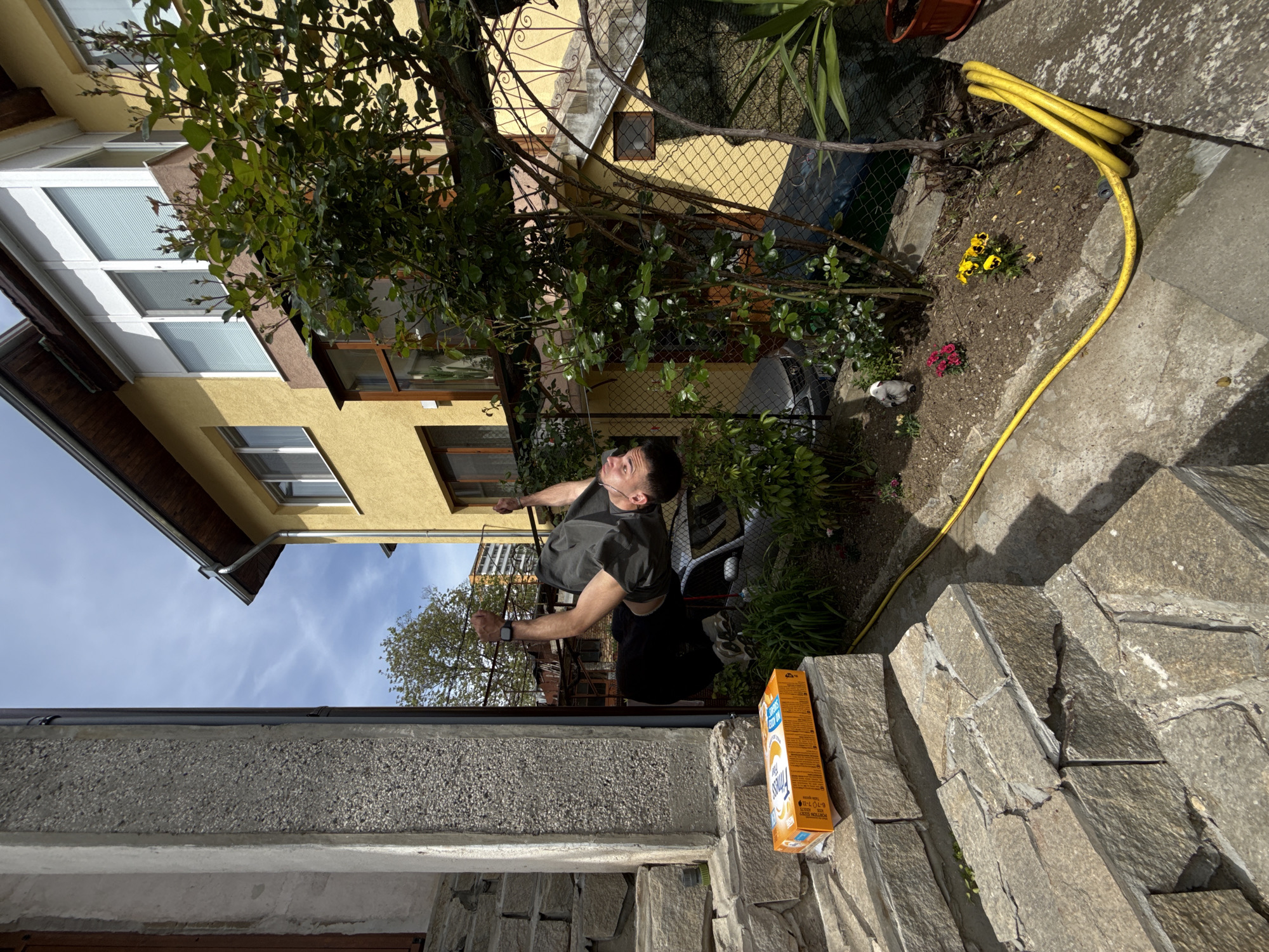 A man in a grey shirt and black pants is stretching his arms in what appears to be a backyard in Shumen, Bulgaria. He is positioned in front of a yellow building and near a garden with a yellow hose. A box of "Fitness" cereal sits on the stone steps of a building. The scene is viewed from a low angle and is rotated counter-clockwise. The sky is visible in the background.
