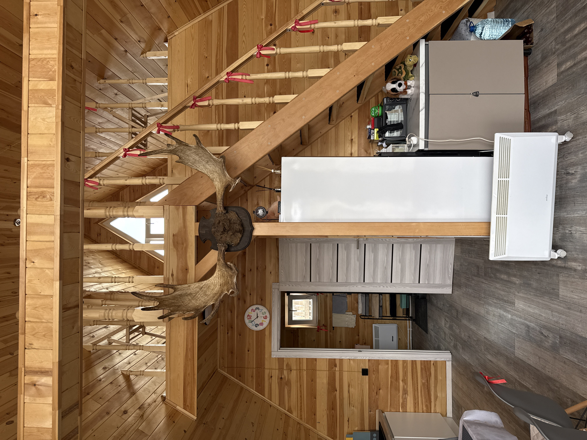 The image shows the interior of a wood-paneled room, with a staircase on the left decorated with red ribbons. A large set of moose antlers is mounted on the wall near the staircase. To the right of the staircase is a kitchen area featuring a gray refrigerator, white countertop, and gray cabinets. A white radiator is positioned near the refrigerator. The floor is covered with dark gray laminate flooring. A mirror reflects a view of clothing on a rack. A clock is mounted on the wall near the mirror. The room appears to be a wooden cabin. There is no evidence to connect the image with Krasnoyarsk, Russia.
