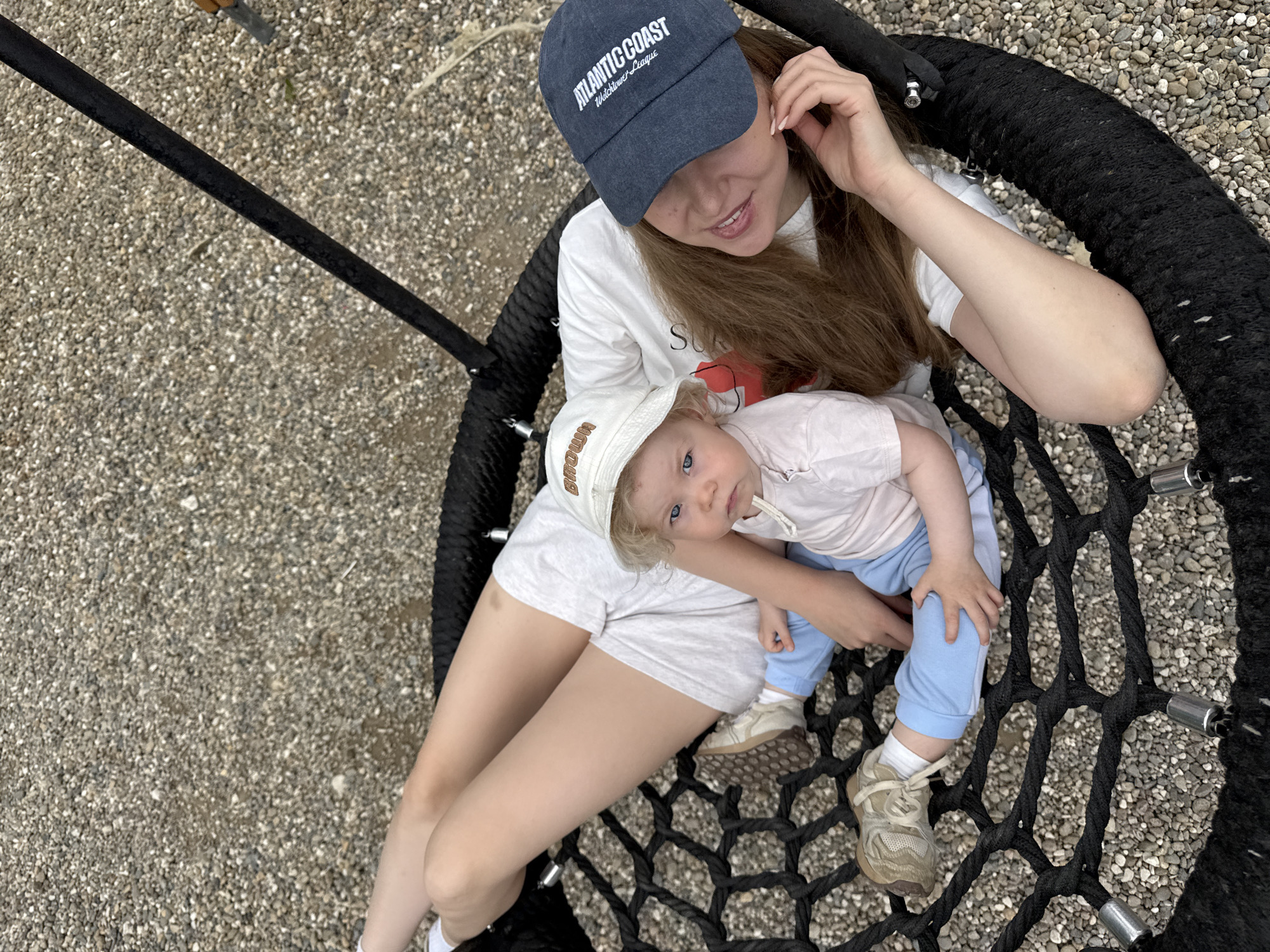 An overhead shot captures a woman and a young child sharing a large, black, rope-mesh nest swing in an outdoor setting. The ground beneath them is covered with light-colored gravel.
The woman, with long brown hair, is wearing a white t-shirt, light-colored shorts, and a dark blue baseball cap. The cap displays white text reading "ATLANTIC COAST" above "Watchtower League." She smiles gently, looking down at the child on her lap, with one hand raised near her ear.
The child, appearing to be a toddler with light-colored hair and blue eyes, sits on the woman's lap, facing the camera. The child wears a light t-shirt, light blue pants, and a cream-colored bucket hat with the word "BROWN" (or "BROWNIE") embroidered on it. Light-colored sneakers are visible on the child's feet, and a hand rests on a knee.
The scene depicts a peaceful and affectionate moment between the two, likely during a leisurely outing at a playground or park in Dobrograd, Russia. The lighting suggests it is daytime, possibly overcast, contributing to a soft, even illumination. The overall mood is calm and content.
