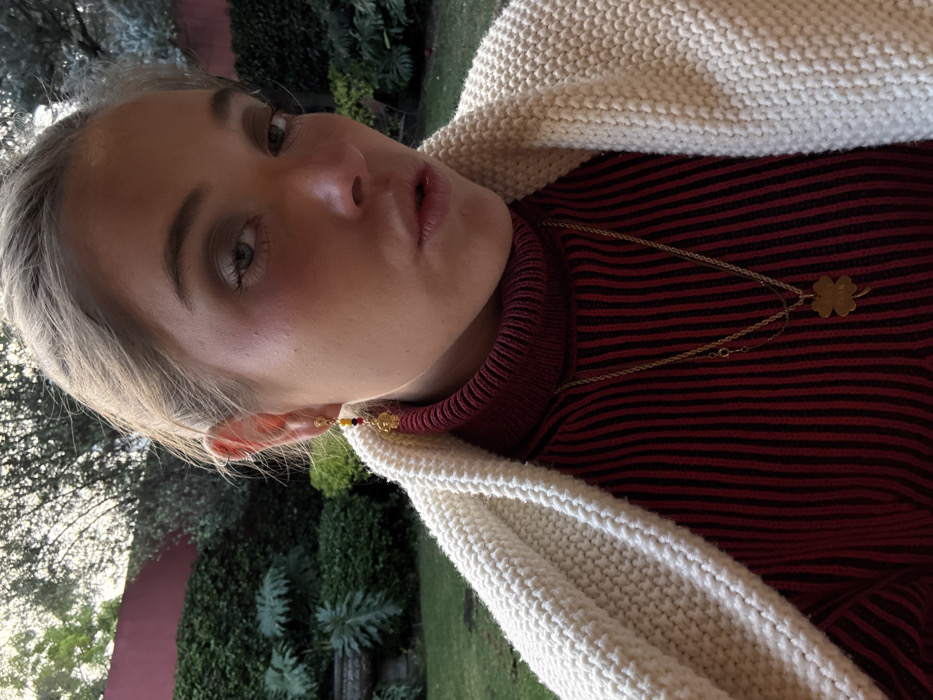 A blonde woman is wearing a red and black striped turtleneck sweater and a white knitted cardigan. She is also wearing a gold necklace with a clover pendant and a beaded earring. The background is blurry, but it appears to be outdoors with trees and greenery. The image is rotated to the left.