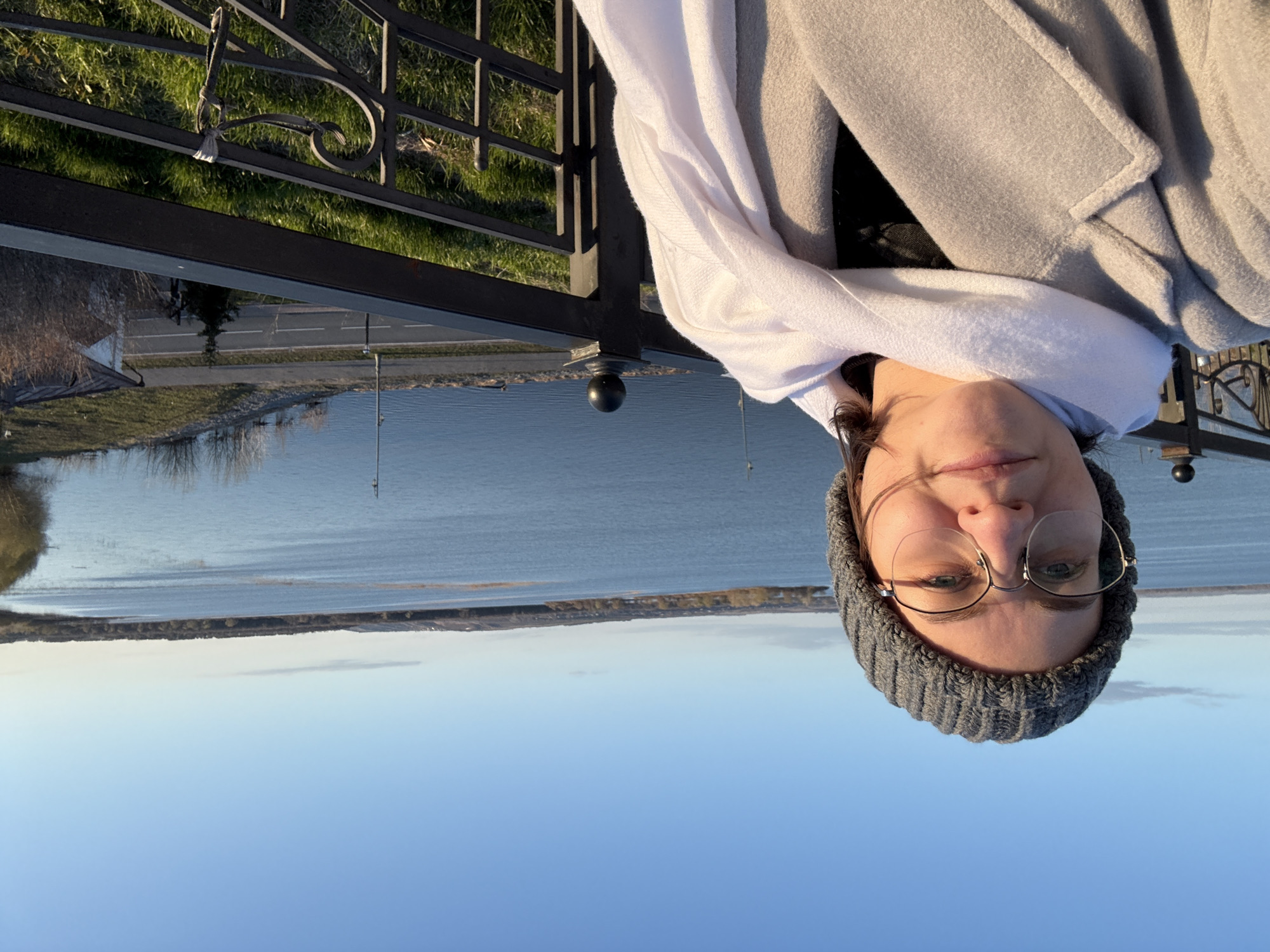 A portrait of a woman wearing a gray beanie, glasses, a white top, and a gray coat. She is standing on a bridge with a metal railing, and behind her is a view of water and land in Sviyazhsk, Russia. The sky is blue.