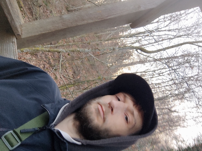 A person wearing a black hoodie and baseball cap poses for a selfie outdoors. Bare trees and foliage are visible in the blurred background. The person is positioned near wooden beams, suggesting a structure like a deck or tower. A green strap or belt is partially visible over their jacket. The light is soft and natural, indicating it might be daytime.