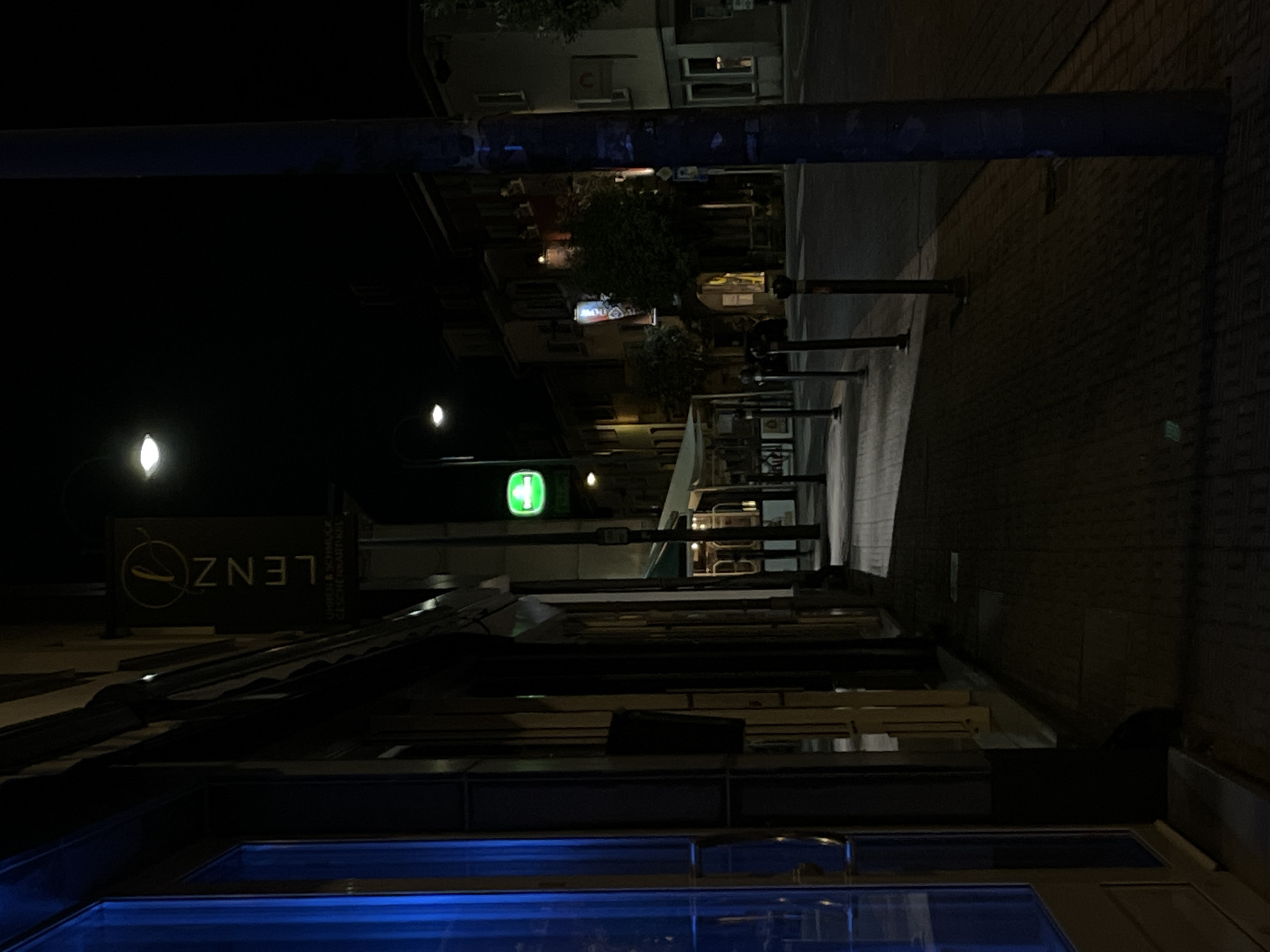 The image captures a nighttime street scene in Saint-Vith, Belgium. A paved sidewalk runs along the left side of the frame, lined with storefronts. One storefront on the left features a large window with a blue tint. Further down the sidewalk, a vertical sign for "LENZ" is visible, accompanied by smaller text indicating "UHREN & SCHMUCK GESCHENKARTIKEL" (Watches & Jewelry Gift Items). A green illuminated sign is also present. The street itself is dark, with the exception of streetlights casting pools of light on the sidewalk and road. Lampposts line the sidewalk, and buildings are visible across the street. The overall atmosphere is quiet and deserted.