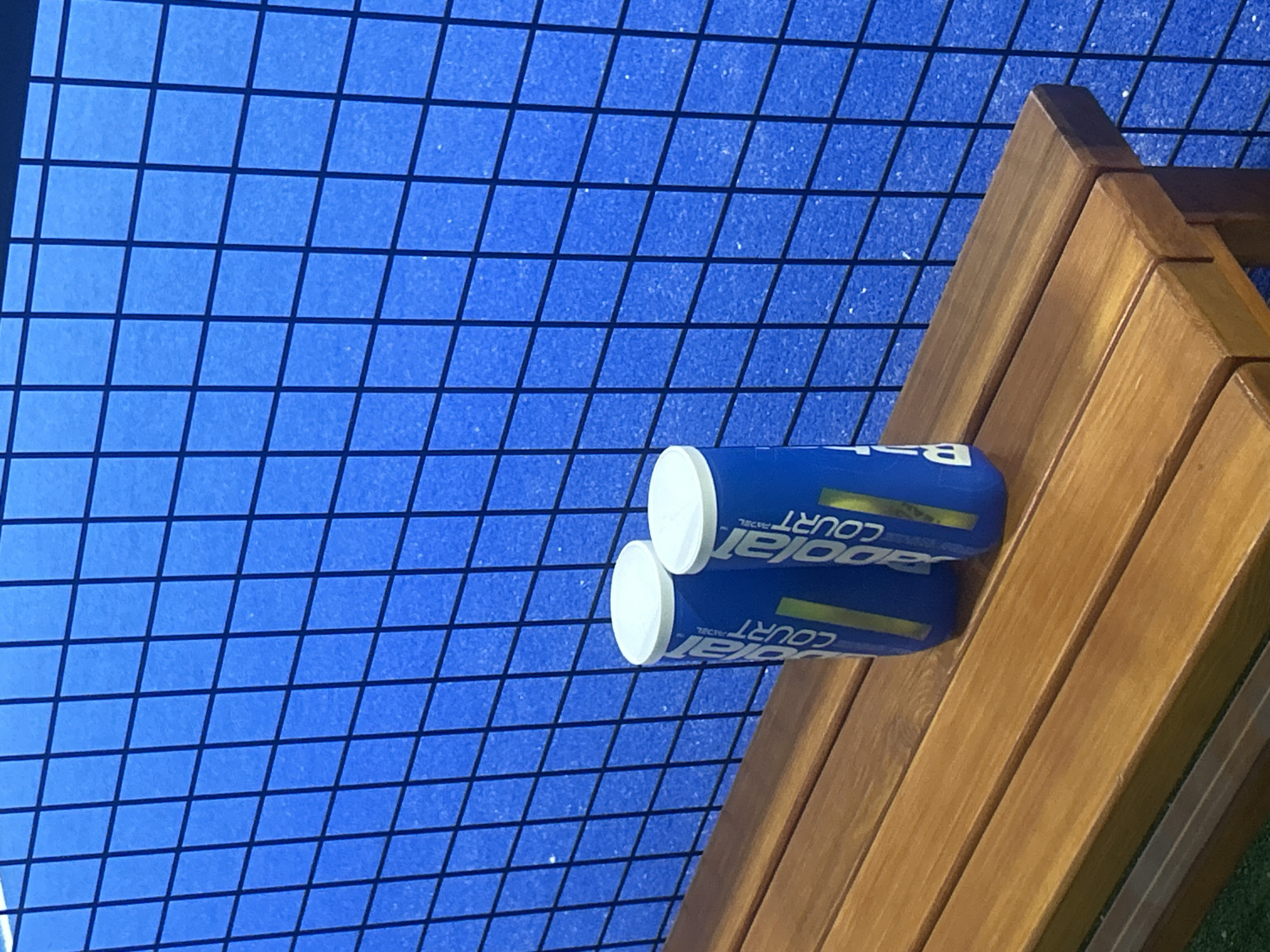 Two cans of Babolat Padel Court tennis balls sit on a wooden bench against a blue, wire-mesh background, likely a tennis court. The scene is in Pleven, Bulgaria.
