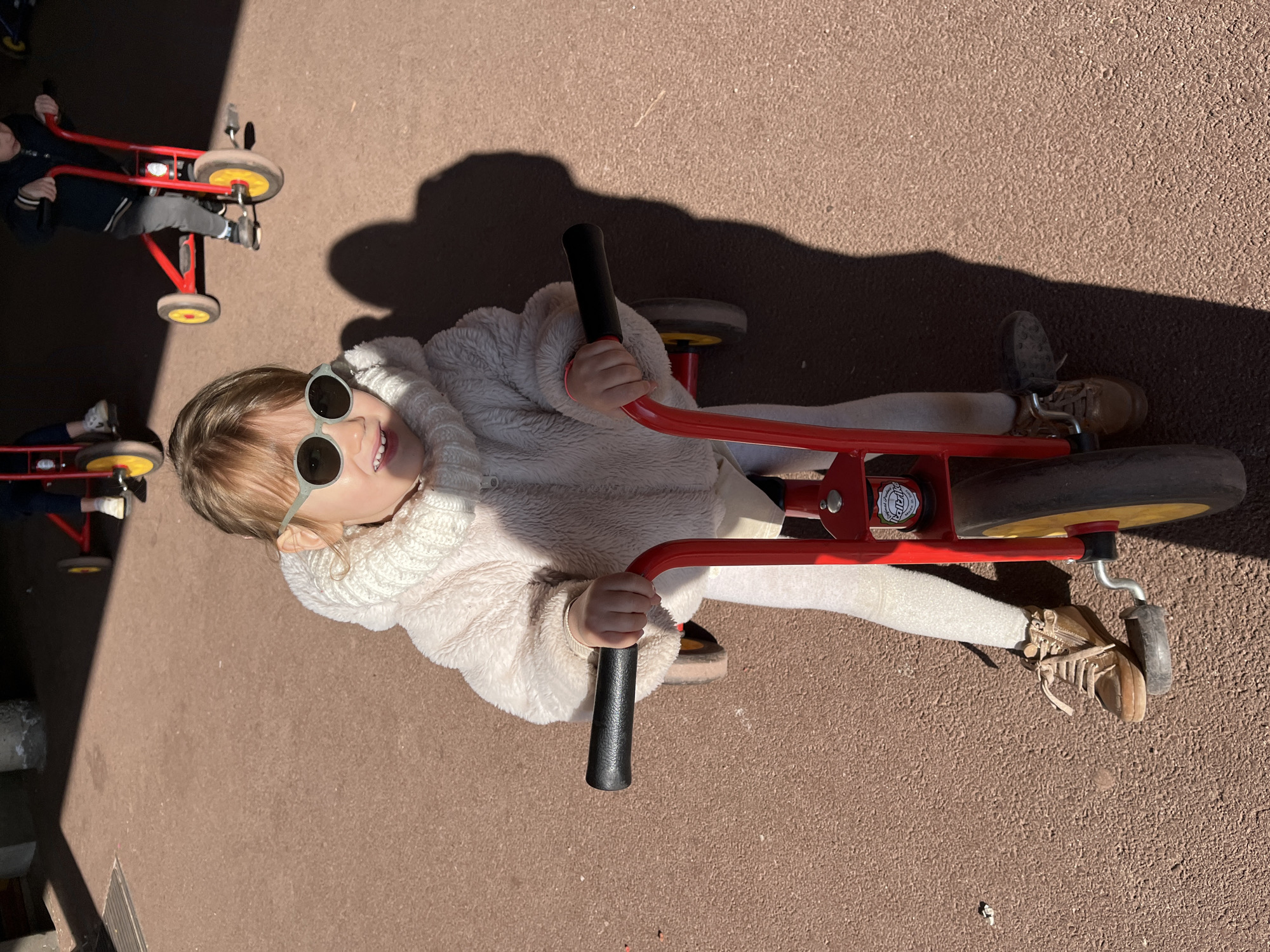 A child wearing sunglasses, a cream-colored fleece jacket, and light-colored pants is riding a red tricycle with black handlebars. The tricycle has yellow wheels. The child is smiling and looking up at the camera. Other children are riding similar tricycles in the background. The scene appears to be outdoors on a reddish-brown surface. The location is La Fouillouse, France.