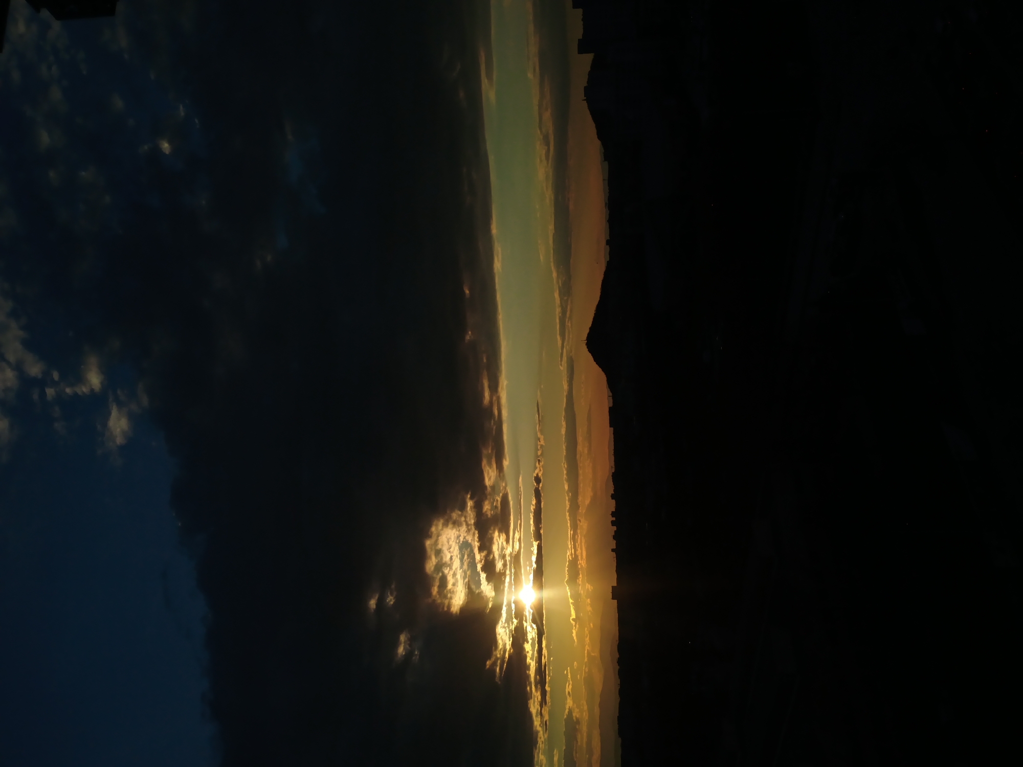 The image shows a view of Plovdiv, Bulgaria, at sunset. The sun is positioned near the center of the frame, with rays of light shining through clouds. The skyline of Plovdiv is visible in silhouette on the right side of the image. The left side features dark clouds, with a hint of blue sky in the upper left corner.