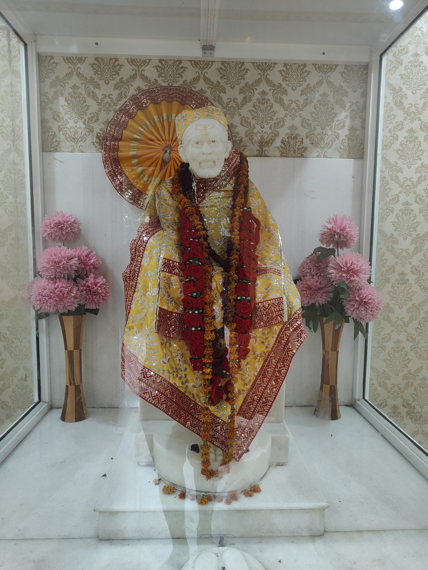 The image depicts a statue of Sai Baba, likely housed in a temple or shrine. The statue is adorned with a yellow and red garment, several garlands of flowers, and a decorative umbrella-like fan behind its head. Pink flowers in wooden vases are placed on either side of the statue. The background features patterned wallpaper and a white wall. The flooring appears to be white marble. The scene is viewed through a glass enclosure, as indicated by reflections. The overall presentation suggests a place of worship or a decorative display in a public space. This scene is located in Gurgaon, India.