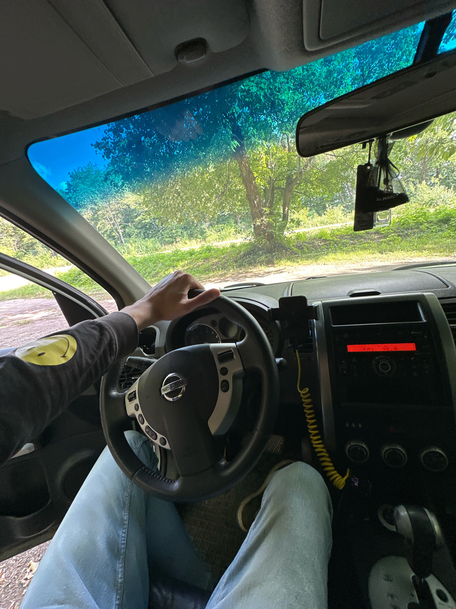 The image shows the interior of a car, viewed from the driver's perspective. A person's hands are on the steering wheel, and their legs are visible, clad in light blue jeans. The dashboard features a Nissan steering wheel, a car stereo displaying "FM1 98.0", and a mobile phone mounted on a holder. Outside the car, through the windshield, a tree-lined landscape with green foliage and sunlight filtering through the leaves is visible, suggesting a rural or park-like setting. A car air freshener is hanging from the rearview mirror. The image was taken in the city of Shatava, Ukraine.