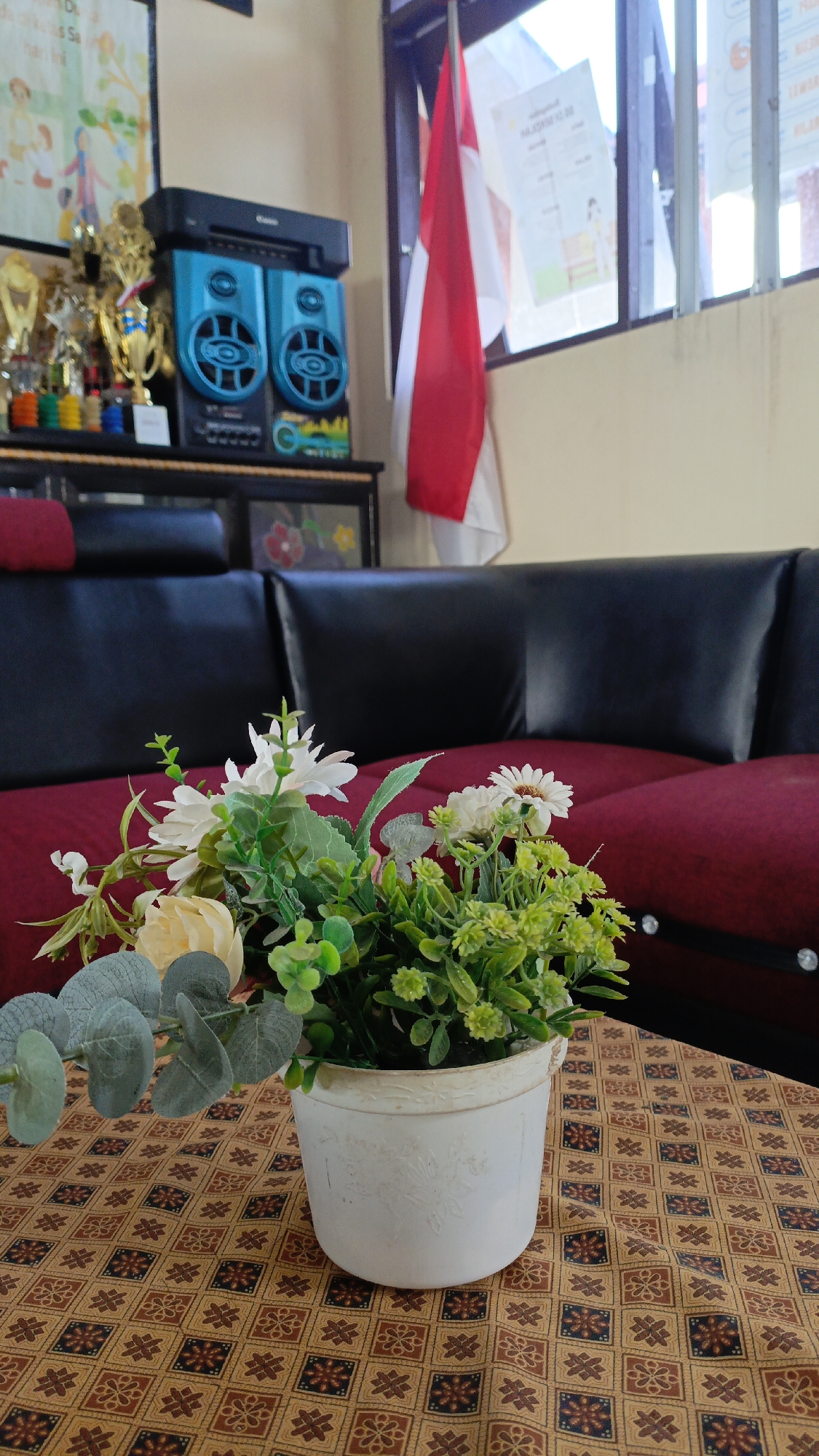 The image depicts an indoor scene, likely a living room, with a focus on a floral arrangement in a white pot placed on a patterned tablecloth. Behind the floral arrangement, a dark-colored sofa with red cushions is visible. In the background, on a shelf, there are trophies, a black and blue stereo system, and a framed picture. A partially visible Indonesian flag is hanging near a window, which reveals a glimpse of the outside. The overall impression is of a domestic setting, possibly in Saring Sungai Binjai, Indonesia, given the presence of the Indonesian flag. No people are present in the image.