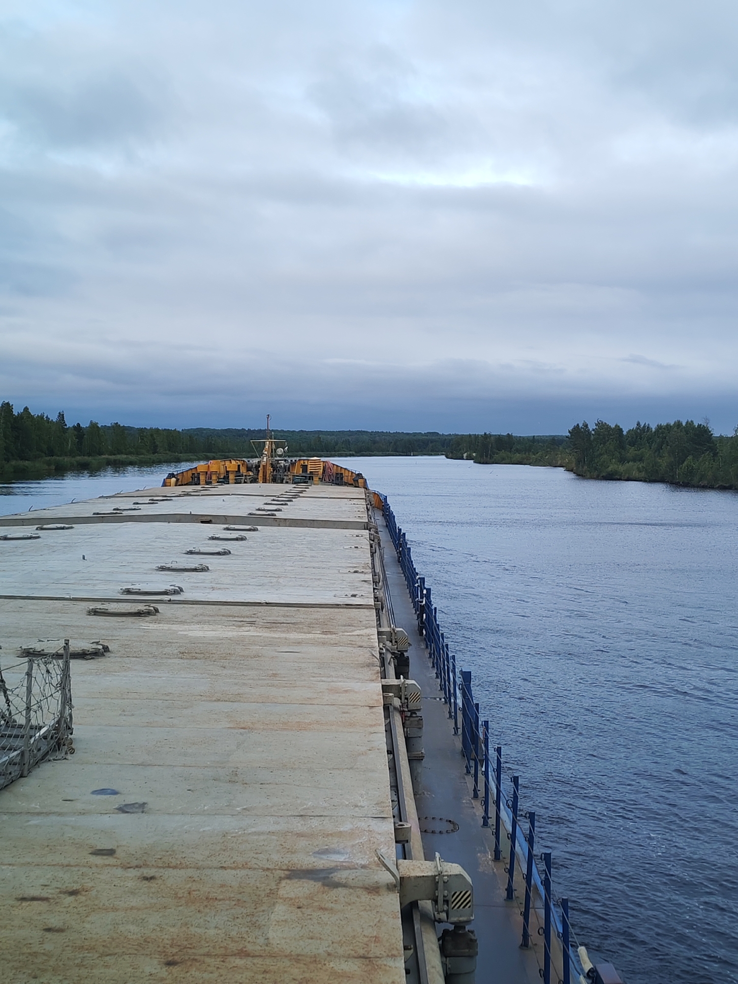 The image depicts a wide, calm river with a barge or cargo ship moving along it. The barge is flat-topped with a textured surface, marked by circular hatches. The barge's stern is visible, featuring yellow equipment and a mast. The riverbanks are lined with dense green trees, suggesting a natural, undeveloped environment. The sky is overcast with pale, diffused light, indicating a cloudy day. The scene appears to be in a rural or semi-rural area, consistent with the location of Ozernoe Ust'e, Russia. No people are visible in the frame. The overall atmosphere is one of quiet transit on a waterway.