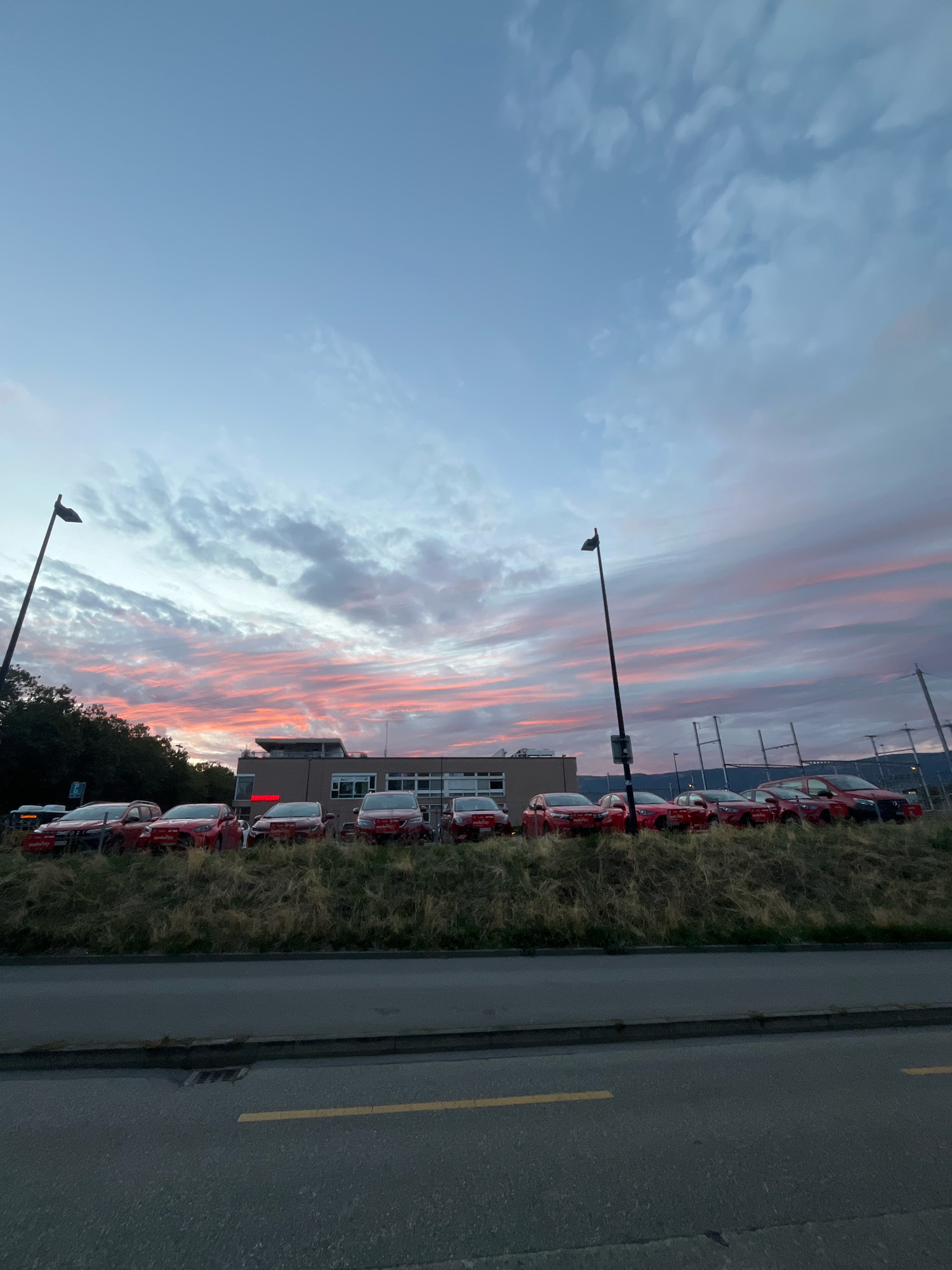 The image captures an outdoor scene at dusk, likely in Yverdon-les-Bains, Switzerland. A line of red cars, possibly a fleet, is parked on a grassy incline in front of a modern-looking building. The sky above is dramatic, with streaks of pink and orange clouds from the setting sun against a backdrop of blue. A street lamp stands tall on the left side of the frame, and another is visible further down the line of cars. The foreground shows a paved road with a yellow dividing line. The overall impression is one of urban infrastructure meeting a colorful natural sky during twilight.