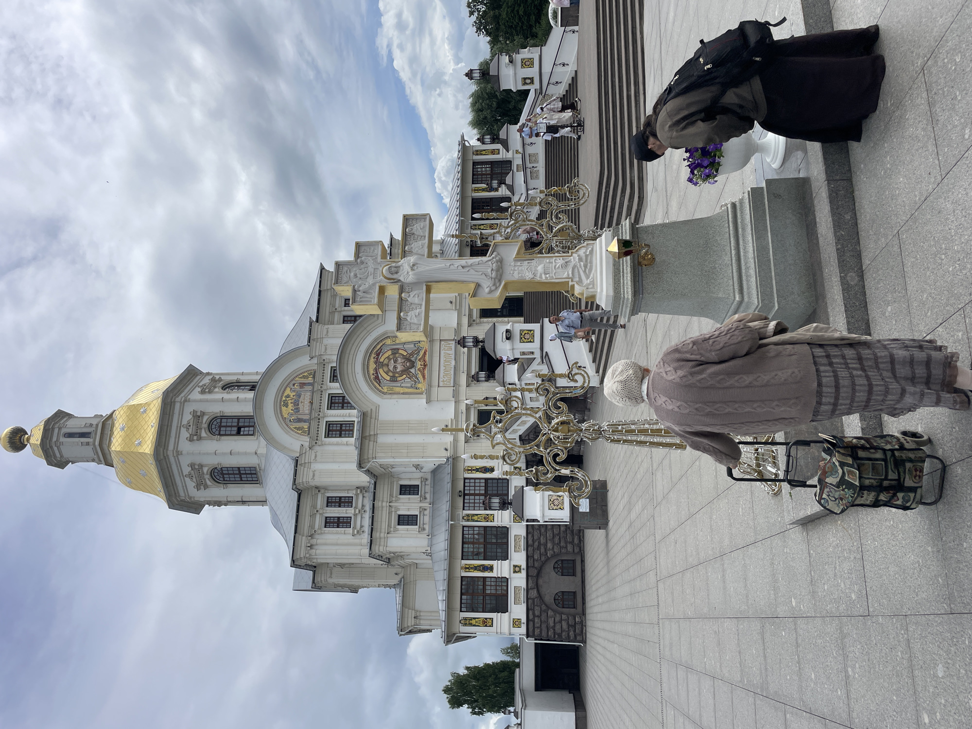 In front of the Holy Trinity Cathedral in Diveyevo, Russia, two women are observed in the foreground. The woman on the left, an elderly individual with white hair, wears a beige knitted sweater and a long striped skirt. She is seen from behind, pushing a patterned rolling shopping cart. To her right, a woman with dark hair and a black beanie bends down, wearing a brown jacket over a long black skirt, and carrying a backpack. She is looking at a white vase filled with purple flowers placed next to a large stone pedestal. This pedestal supports an ornate, gold-trimmed cross with relief sculptures. The cathedral, a grand white structure with a golden dome and mosaics, serves as the backdrop. Several people are visible on the steps leading up to the cathedral entrance. The scene unfolds on a paved plaza under a cloudy sky.
