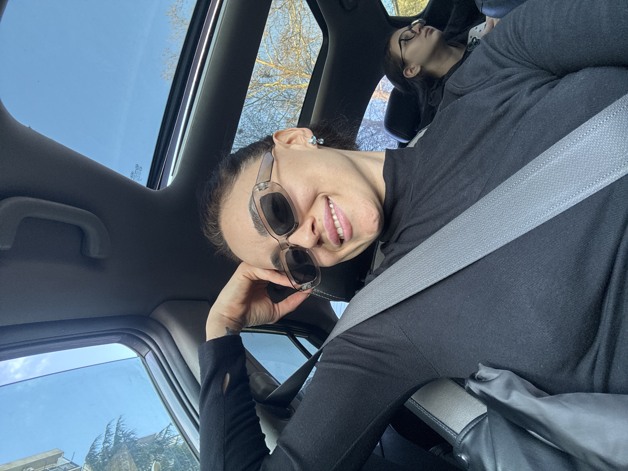 The image depicts a selfie taken from the front passenger seat of a vehicle during daylight hours. A woman, estimated to be in her late 20s to early 30s, is the primary subject. She has dark hair pulled back, wears light-framed rectangular sunglasses, and a black high-necked long-sleeved top. A grey seatbelt is fastened diagonally across her chest. Her left hand, with a small dark tattoo visible on the inner wrist, is positioned near her temple, with fingers slightly curled. She is smiling directly at the camera. She wears small blue, flower-shaped earrings.
In the background, a second individual, appearing to be a younger female with dark hair and round eyeglasses, is partially visible in the rear passenger seat, looking towards the right side of the vehicle. A black garment or bag rests on the primary subject's lap. The vehicle's interior is dark. Through the panoramic sunroof, a clear blue sky and the bare branches of deciduous trees are visible. Through the side window, a light-colored multi-story residential building with balconies and the foliage of evergreen trees are discernible. The overall environment suggests an urban or suburban residential area consistent with locations found in Tbilisi, Georgia.