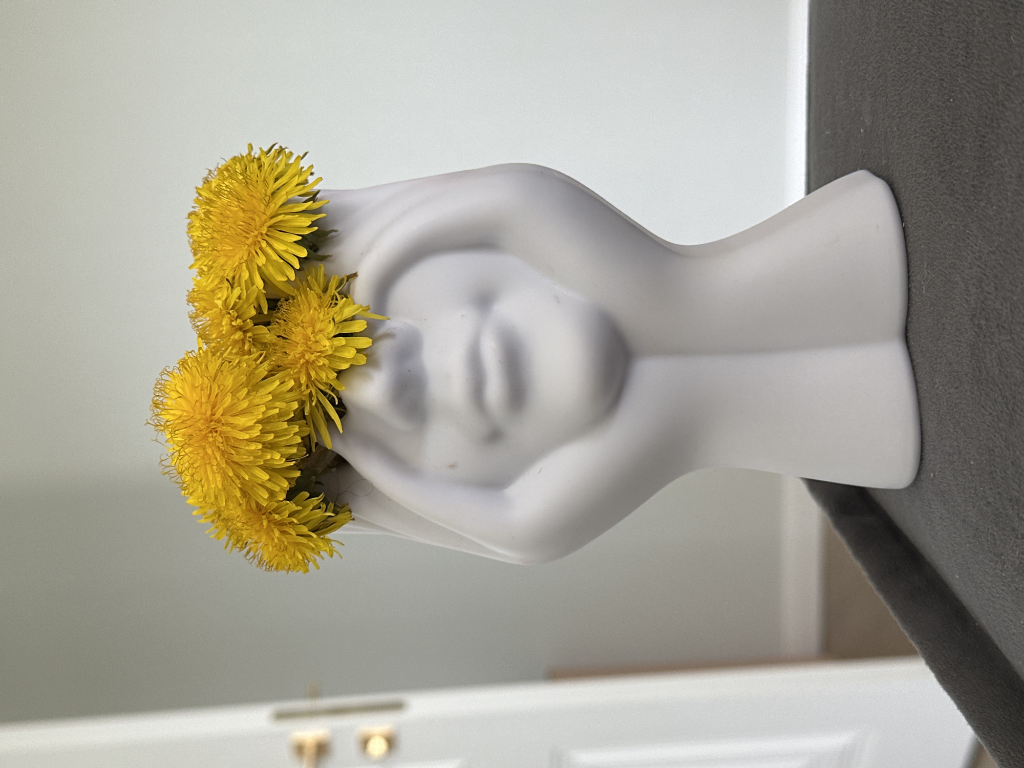 A still life photograph depicts a light gray, sculptural vase with a stylized human face and hands cradling the head. The vase is filled with vibrant yellow dandelions. The vase rests on a dark gray surface, likely a table or countertop. The background is a blurred combination of white walls and a glimpse of a white door with golden hardware. The image is taken indoors. There is no information in the image to confirm the location of Kharnavy, Russia.