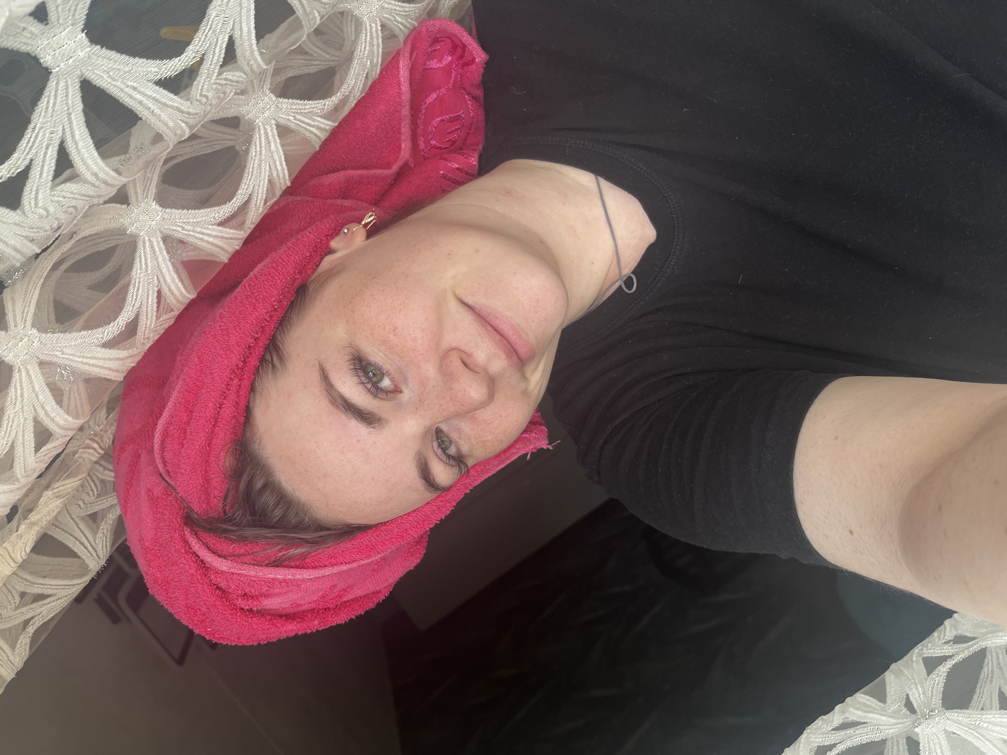 A person is lying down with their head wrapped in a pink towel. They are wearing a black shirt. Part of a white, patterned textile can be seen on the left side of the frame. The location is not discernible from the image.