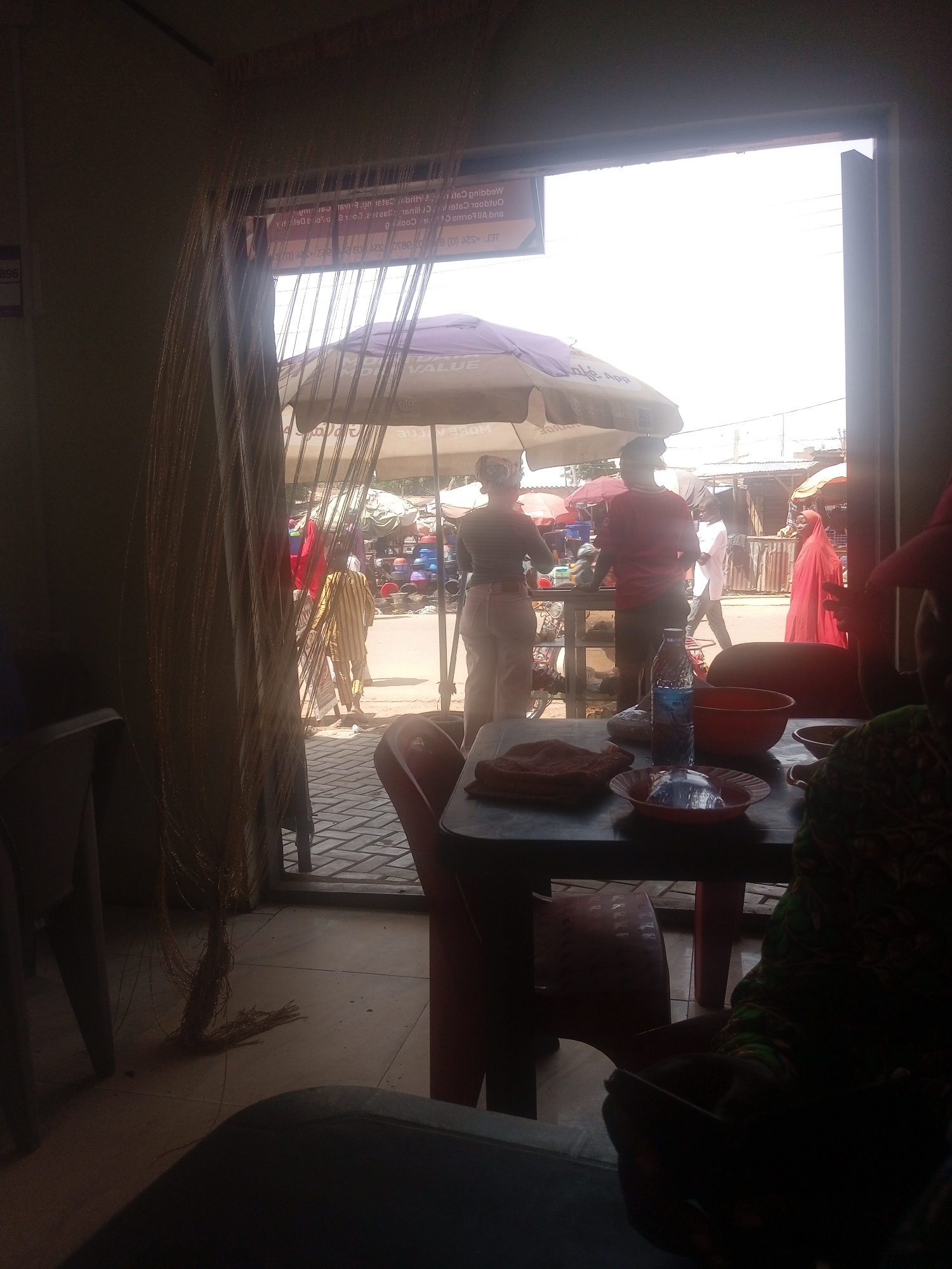 This is a medium-shot, indoor-outdoor photograph taken from within a building looking out into a bustling marketplace. The foreground is dominated by a dark table laden with food items and drinks, and out-of-focus figures seated at it. In the mid-ground, several individuals are seen interacting with vendors under umbrellas. The background reveals a lively street scene with stalls and buildings. The lighting suggests a bright, sunny day. The composition highlights the contrast between the calm interior and the vibrant activity of the marketplace.