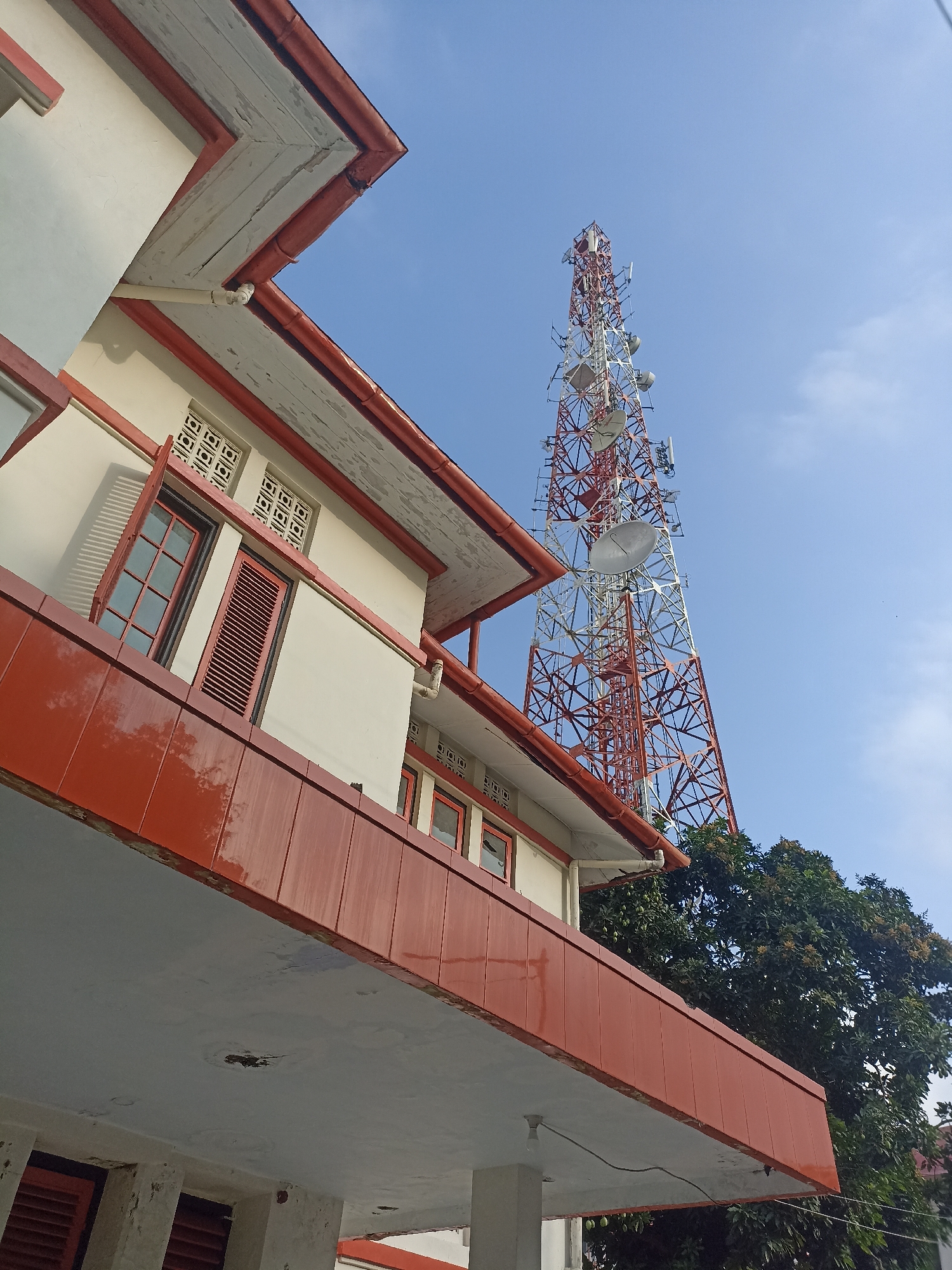 An eye-level, medium shot captures a telecommunications tower against a bright blue sky in Kelurahan Kepatihan, Indonesia. The tower, predominantly red and white, is a lattice structure adorned with various antennas and dishes. In the foreground, a building with a white facade and orange trim features multiple windows, some with louvred shutters. A tree with green foliage is visible to the right of the tower. The image appears to be taken during the day, with the sun casting shadows.